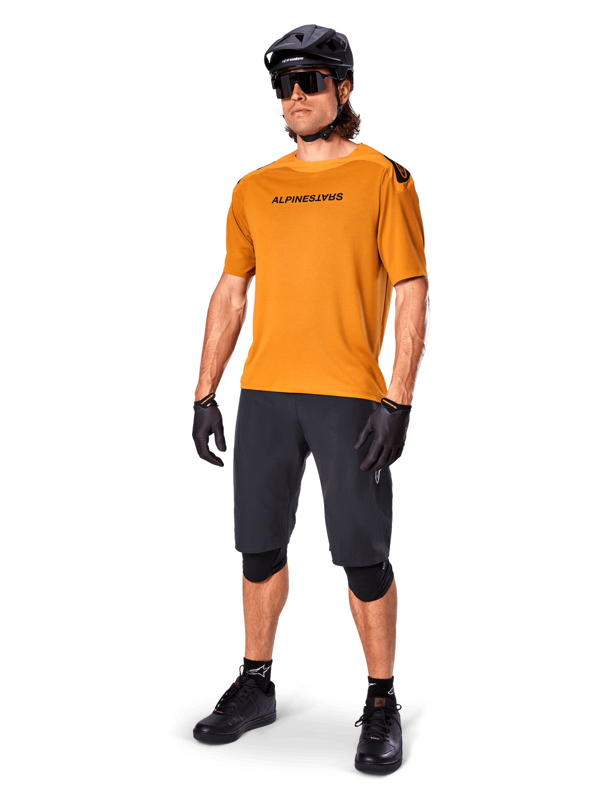 Manlig cyklist i Alpinestars A-Aria Polartec Switch-tröja i Dark Gold, kortärmad MTB-tröja, kombinerad med svarta cykelshorts, knäskydd, svart hjälm och handskar, professionell mountainbike-outfit för stigcykling