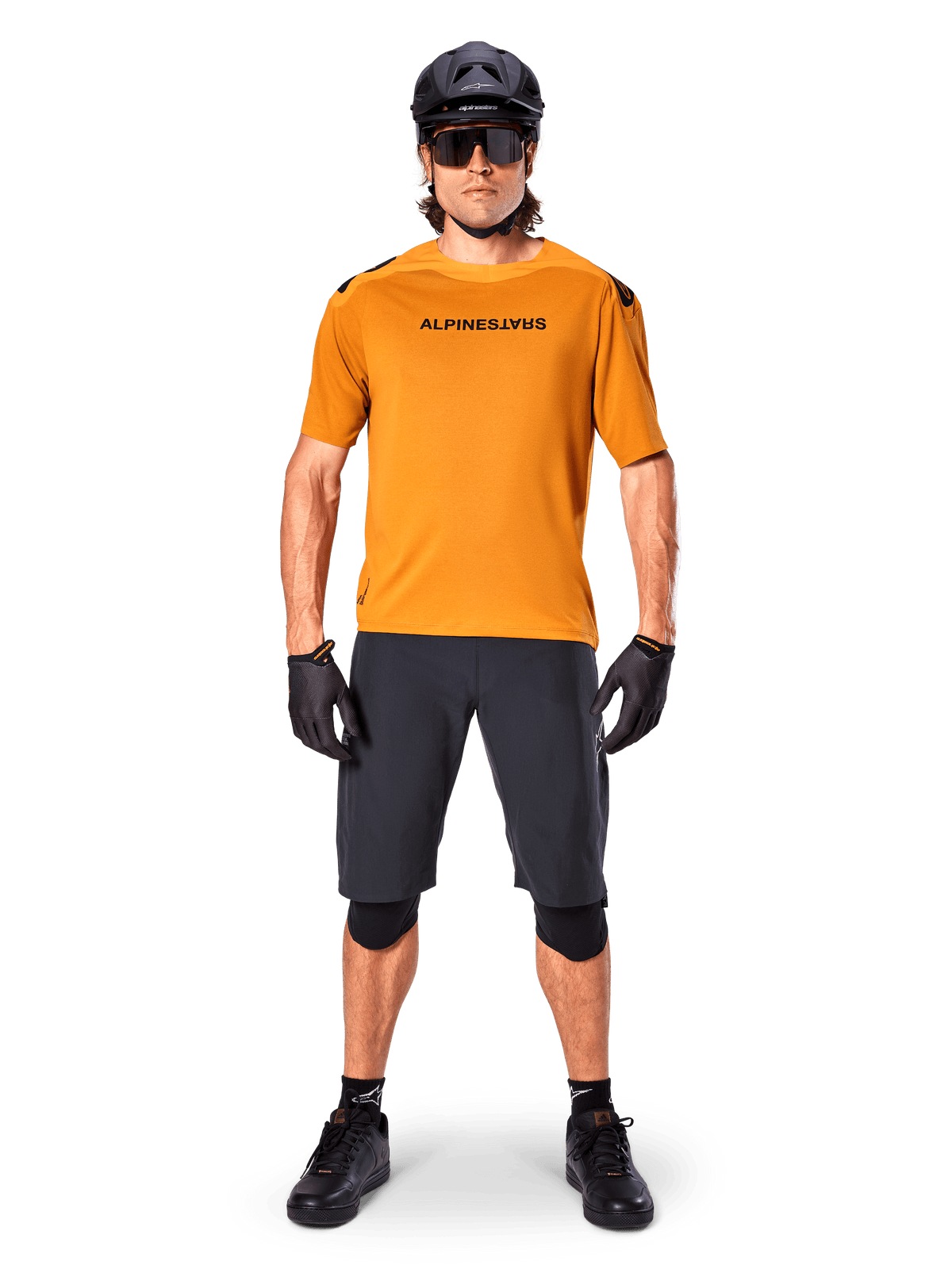 Manlig cyklist bär Alpinestars A-Aria Polartec Switch kortärmad tröja i Dark Gold, kombinerat med svarta MTB-shorts, hjälm, solglasögon och handskar, professionell mountainbike-utrustning
