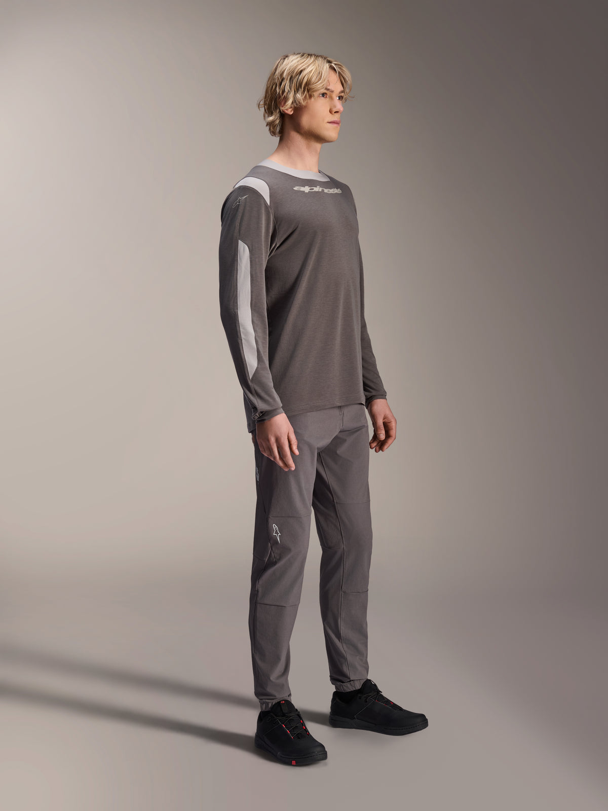 Manlig modell iklädd Alpinestars A-Dura Dri Wool långärmad tröja i mörkgrått, kombinerad med matchande gråa byxor och svarta skor, ljusgrå meshpaneler på axlar och ärmar, teknisk MTB-utrustning för terrängcykling