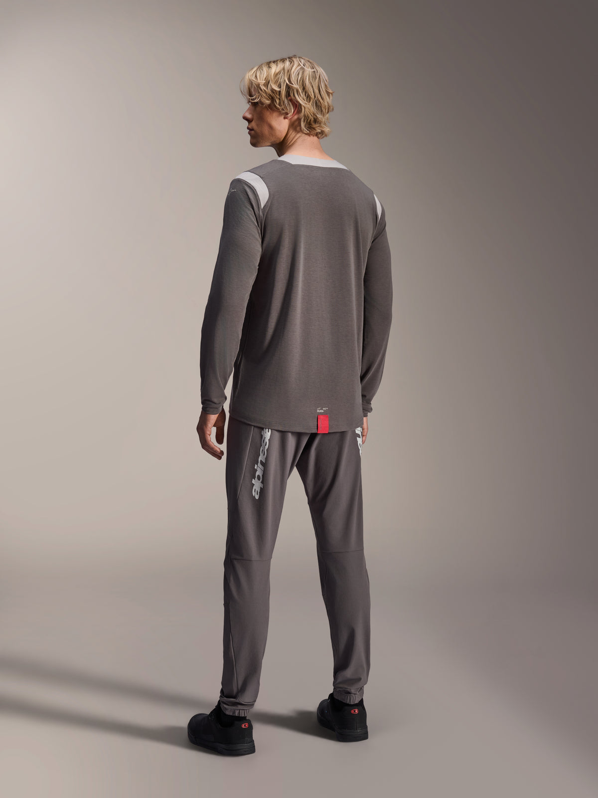 Manlig modell som bär Alpinestars A-Dura Dri Wool Long Sleeve Jersey, mörkgrå med ljusgrå meshpaneler på axlarna, matchande mörkgrå cykelbyxor, vy bakifrån som visar det längre bakstycket, mountainbike-kläder