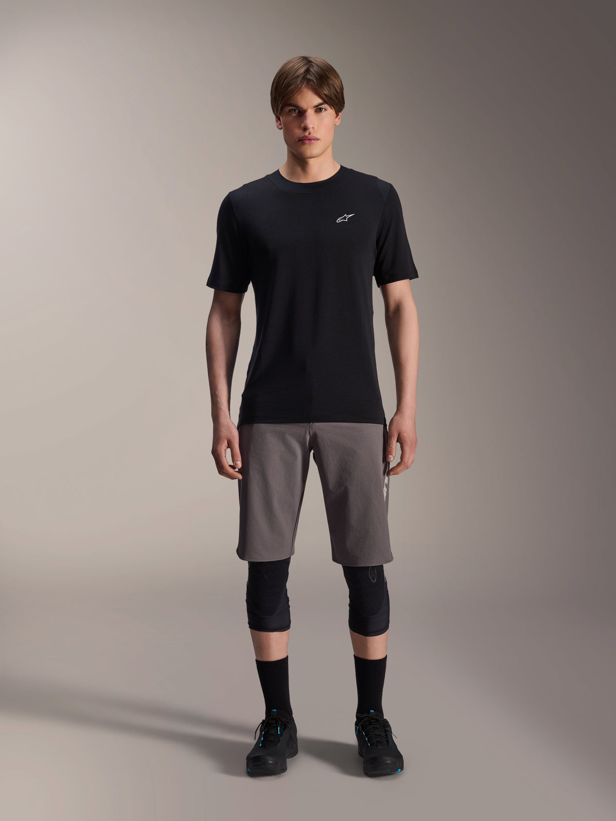 Alpinestars A-Dura Dri Wool kortärmad tröja, svart, bärs av en modell med grå mountainbikeshorts, svarta knäskydd och svarta cykelskor, minimalistisk design med en liten vit Astars-logotyp på bröstet, funktionella MTB-kläder för stigcykling