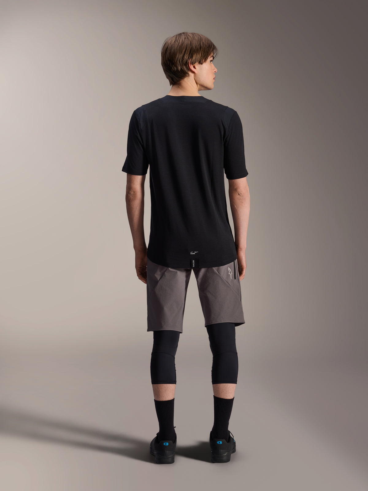 Alpinestars A-Dura Dri Wool kortärmad tröja, svart, sedd bakifrån på en modell med gråa MTB-shorts och svarta knäskydd, med ett förlängt bakstycke och atletisk passform för terrängcykling