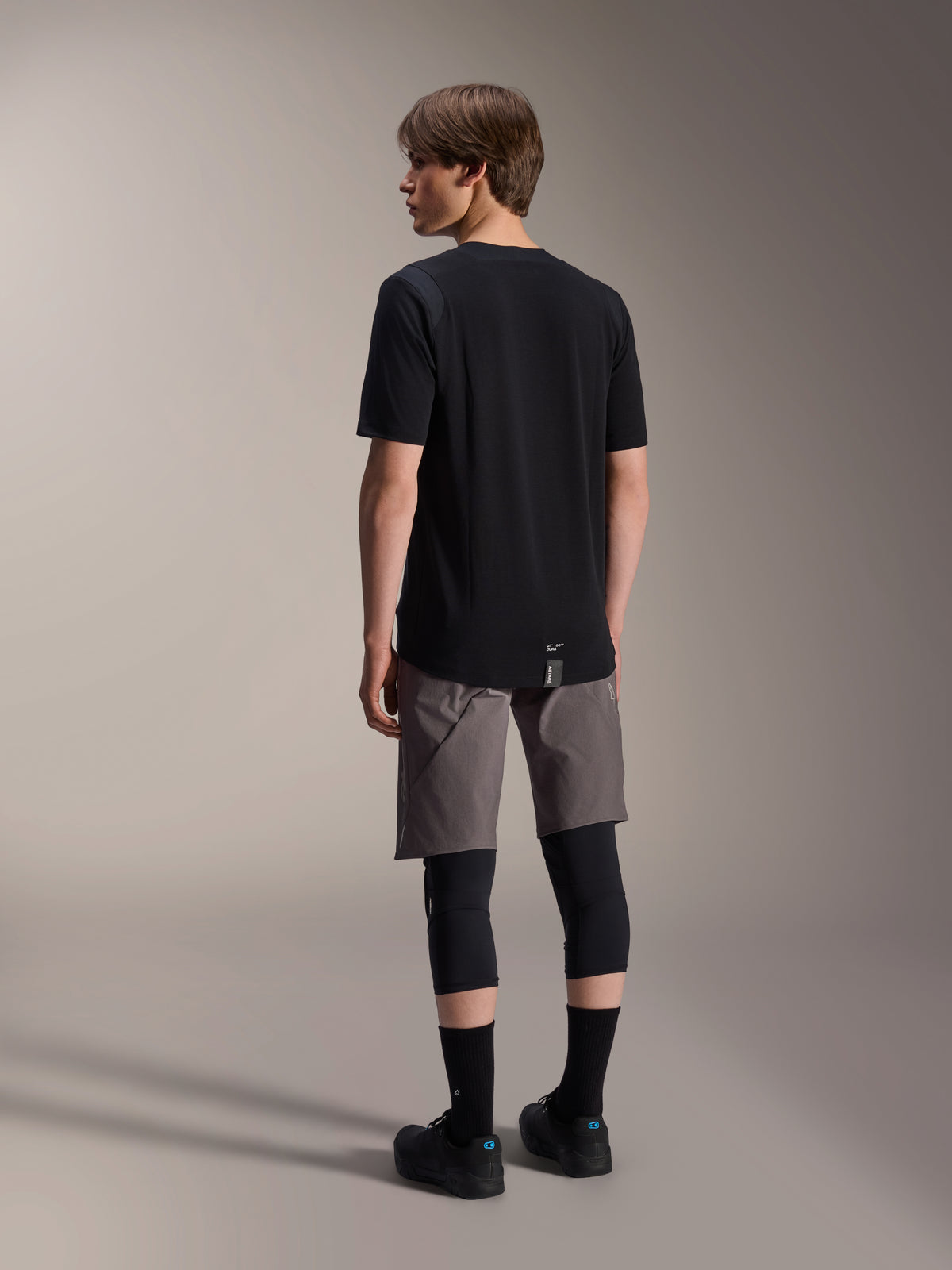 Alpinestars A-Dura Dri Wool kortärmad tröja, svart, ryggvy på modell, bärs med grå mountainbike-shorts och svarta knäskydd, med förlängt bakstycke och en diskret logotyp, tekniska MTB-kläder
