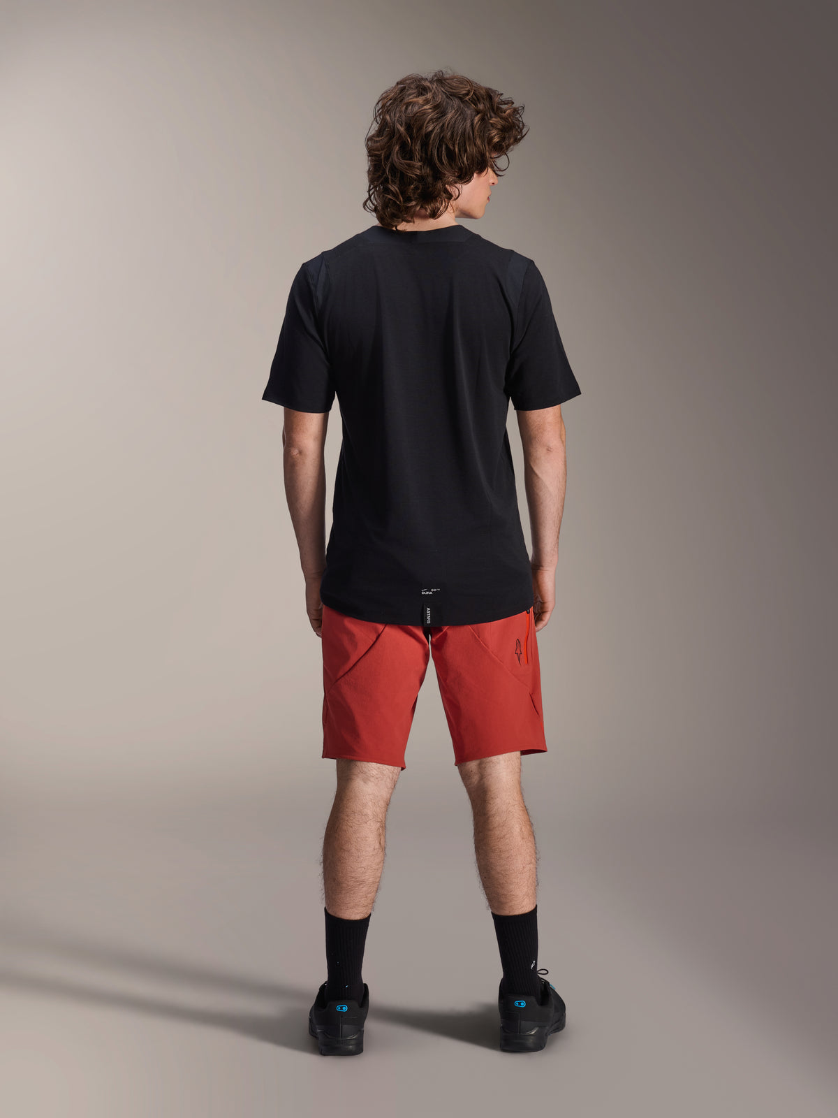 Manlig modell sedd bakifrån i Alpinestars A-Dura Elite-shorts i Dark Orange och en svart kortärmad A-Dura Dri Wool-tröja, svarta strumpor och cykelskor, professionella mountainbikekläder