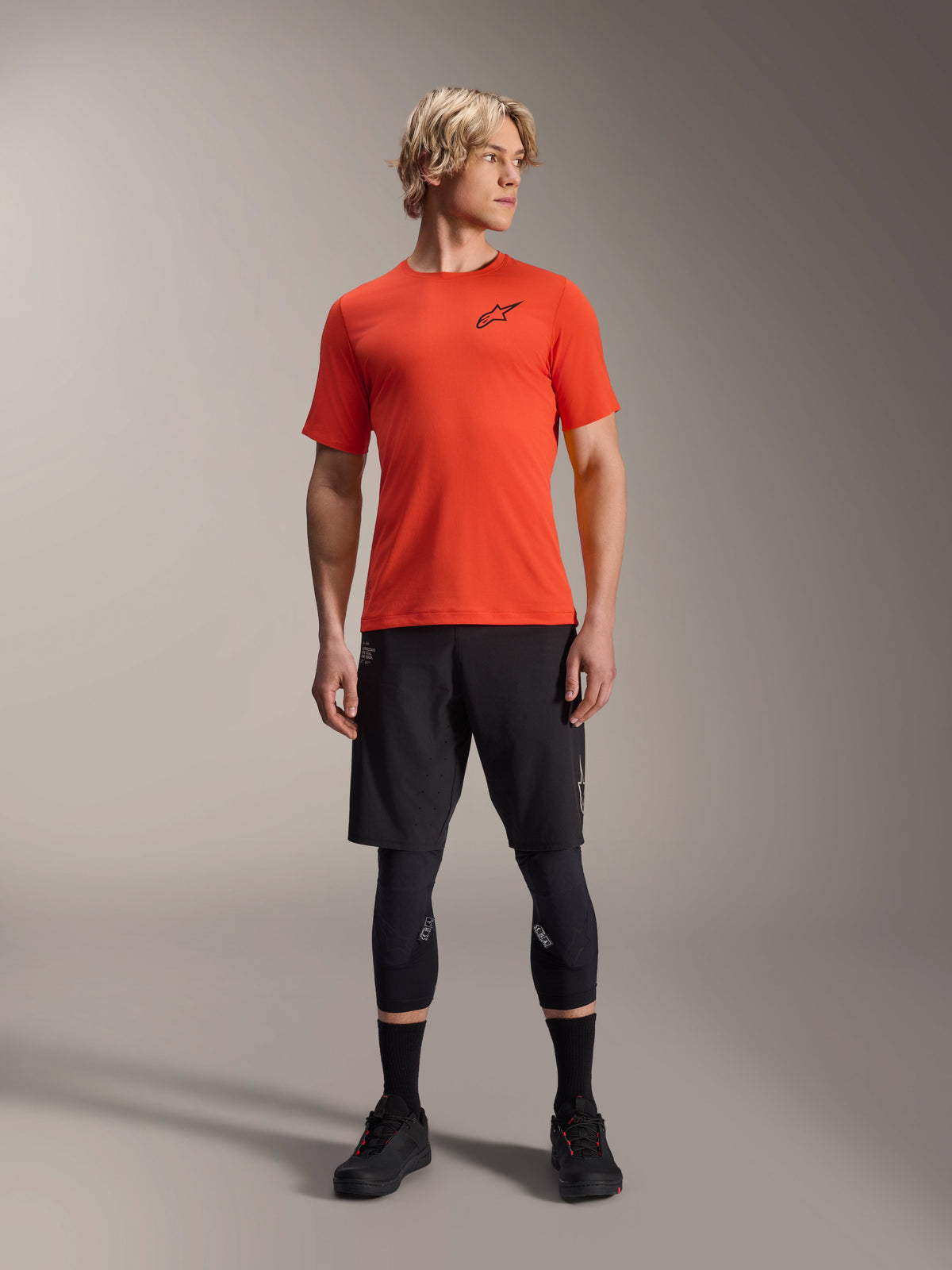 Alpinestars A-Aria Astar kortärmad tröja, Spicy Orange, buren av modell med svarta mountainbikeshorts och knäskydd, lätt och andningsbart prestandaplagg för MTB