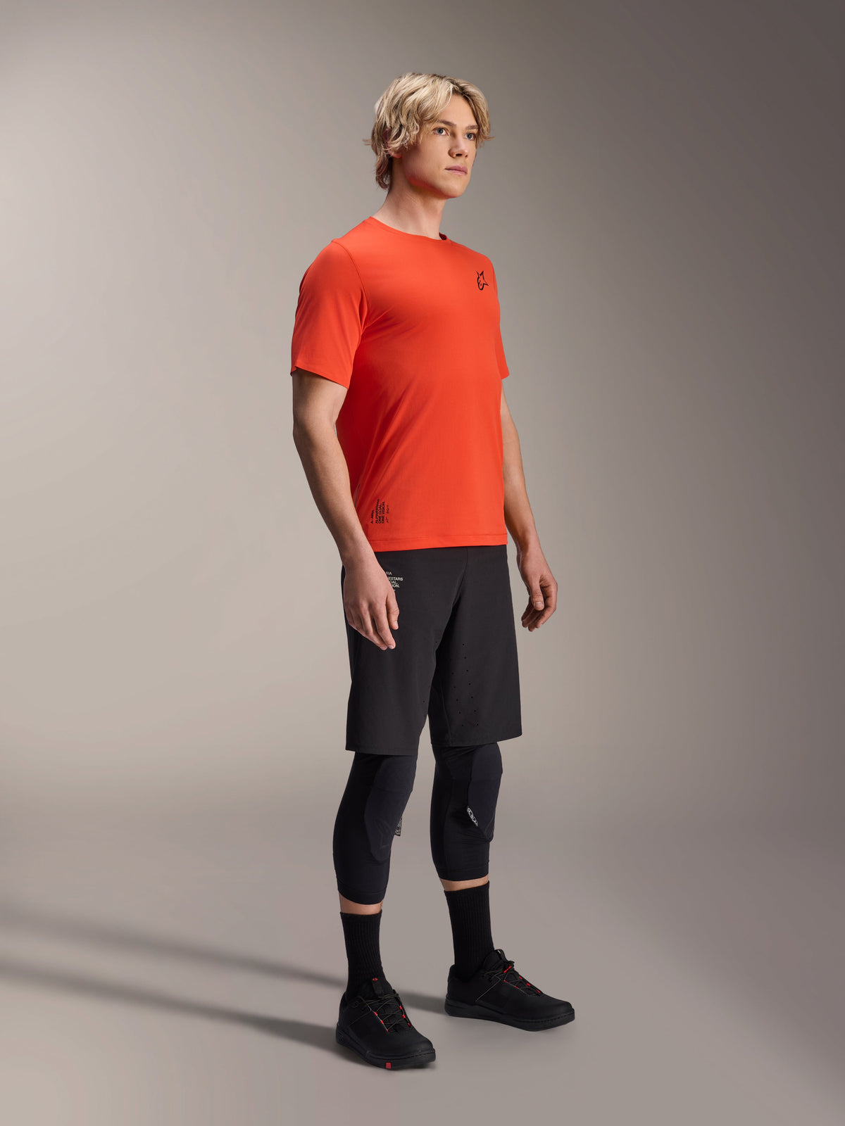 Alpinestars A-Aria Astar kortärmad tröja i Spicy Orange, bärs av en modell med svarta MTB-shorts och knäskydd, lätta och ventilerande mountainbike-kläder, livfull orange med minimal svart logotyp