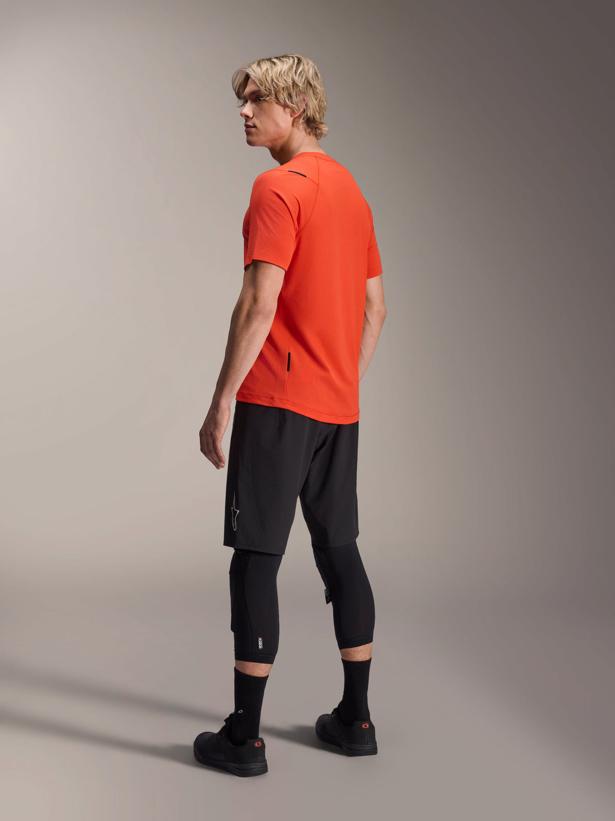 Alpinestars A-Aria Astar kortärmad tröja, Spicy Orange, trekvartsvy bakifrån på modell som blickar över axeln, kombinerad med svarta MTB-shorts och knäskydd, prestationsinriktade mountainbike-kläder