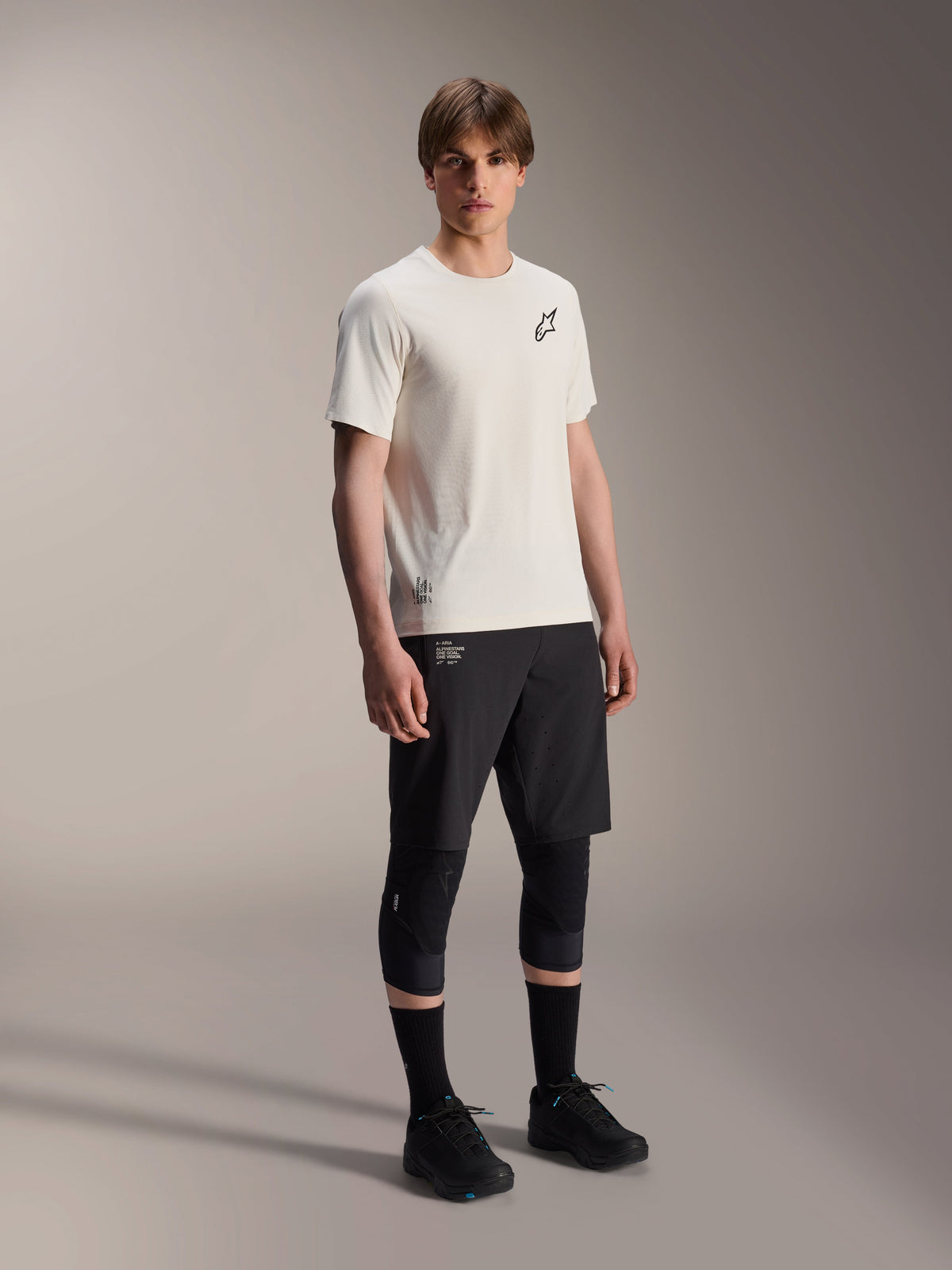 Alpinestars A-Aria Astar tröja, kortärmad, Ecru, med svart bröstlogotyp, bärs med svarta cykelshorts och knäskydd, lättviktig MTB-prestandautrustning