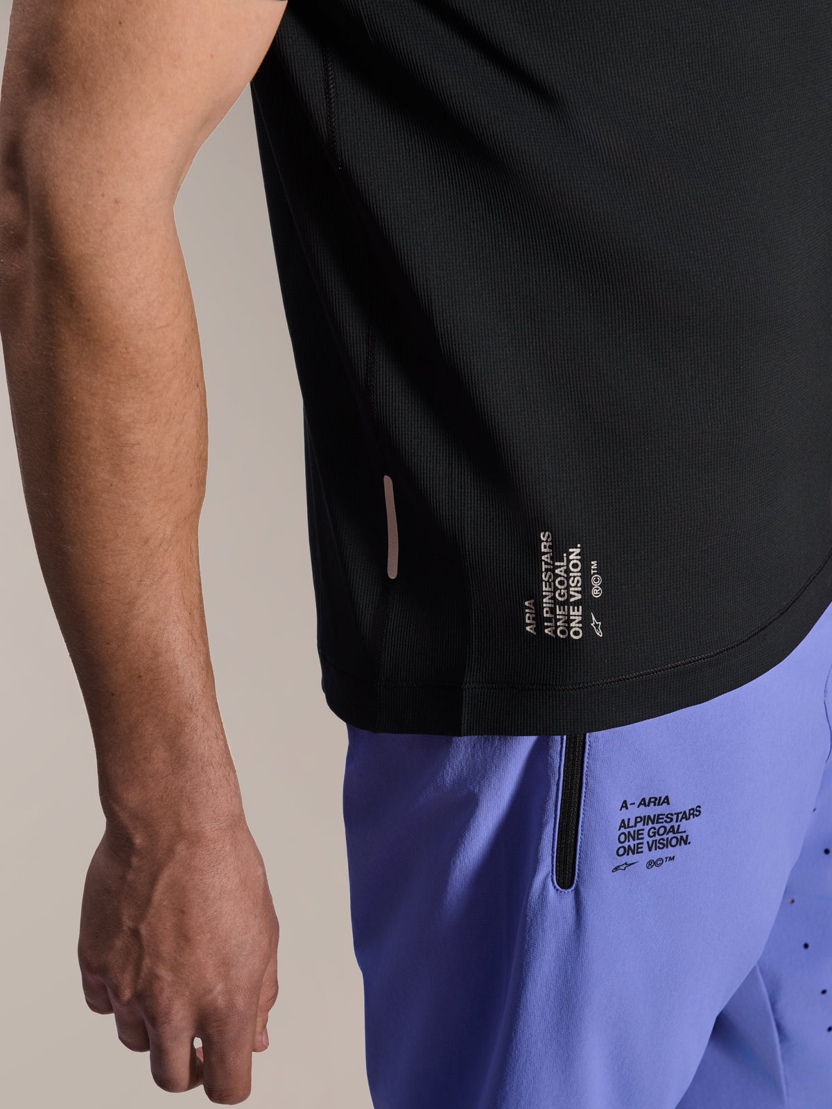 Alpinestars A-Aria Switch Short Sleeve Jersey, Black MTB jersey, close-up of side hem with white 'ONE GOAL. ONE VISION.' branding, worn with light purple cycling shorts featuring matching logos