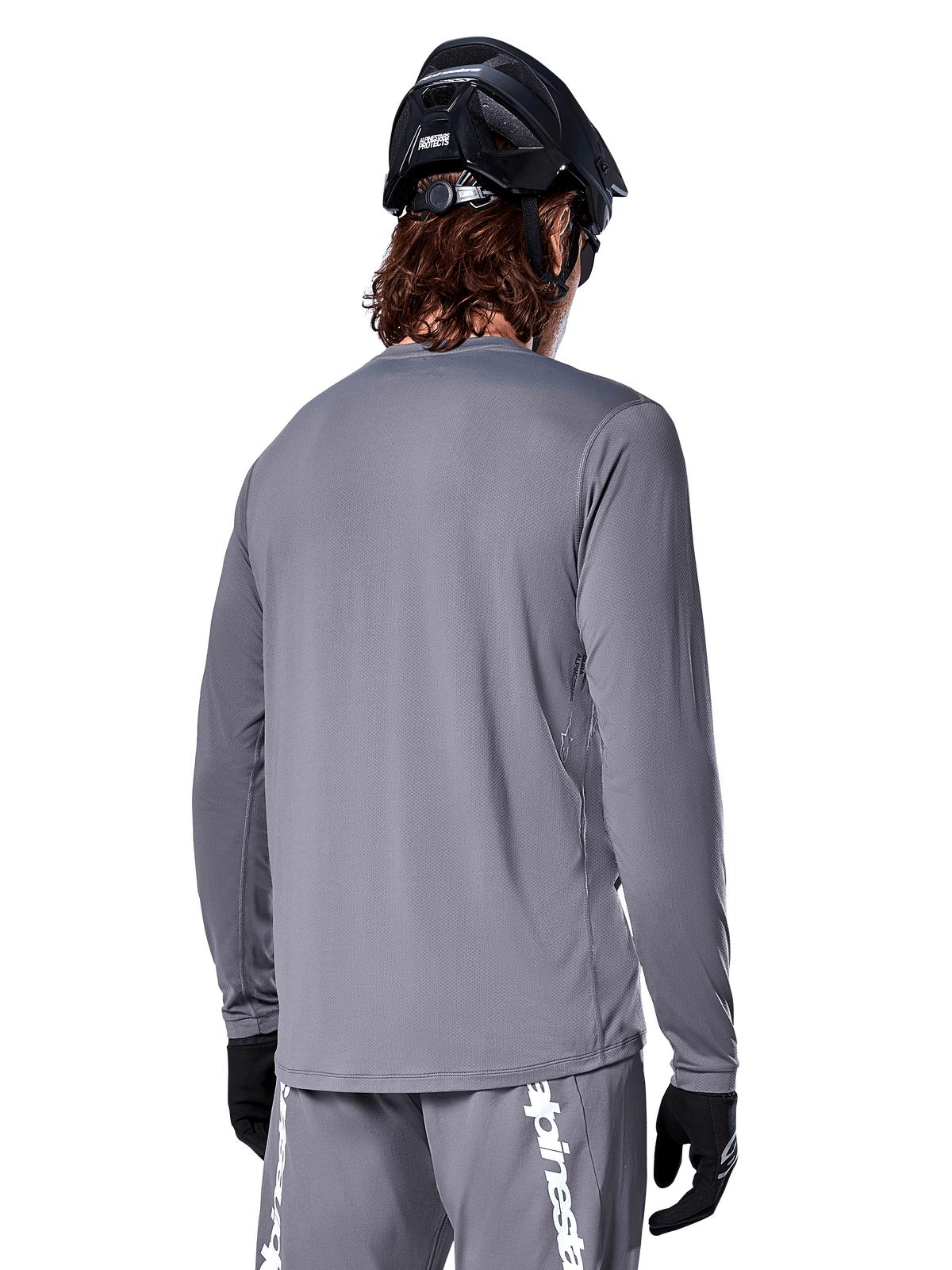 Alpinestars A-Dura Rocker långärmad tröja i mörkgrått, vy bakifrån av mountainbike-cyklist, tillsammans med matchande grå shorts med vit logotyp, svart MTB-hjälm och svarta handskar, professionell utrustning för terrängcykling.
