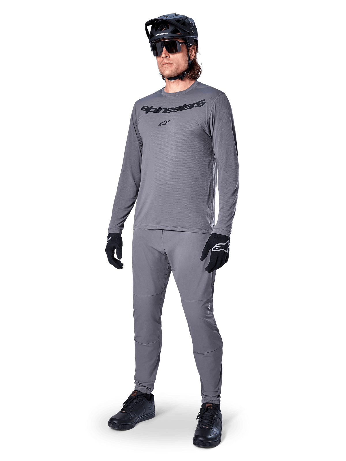 Alpinestars A-Dura Rocker långärmad tröja, mörkgrå, bärs av en modell med matchande grå byxor, svart mountainbikehjälm, solglasögon och handskar, avslappnad MTB-stil för terrängkörning