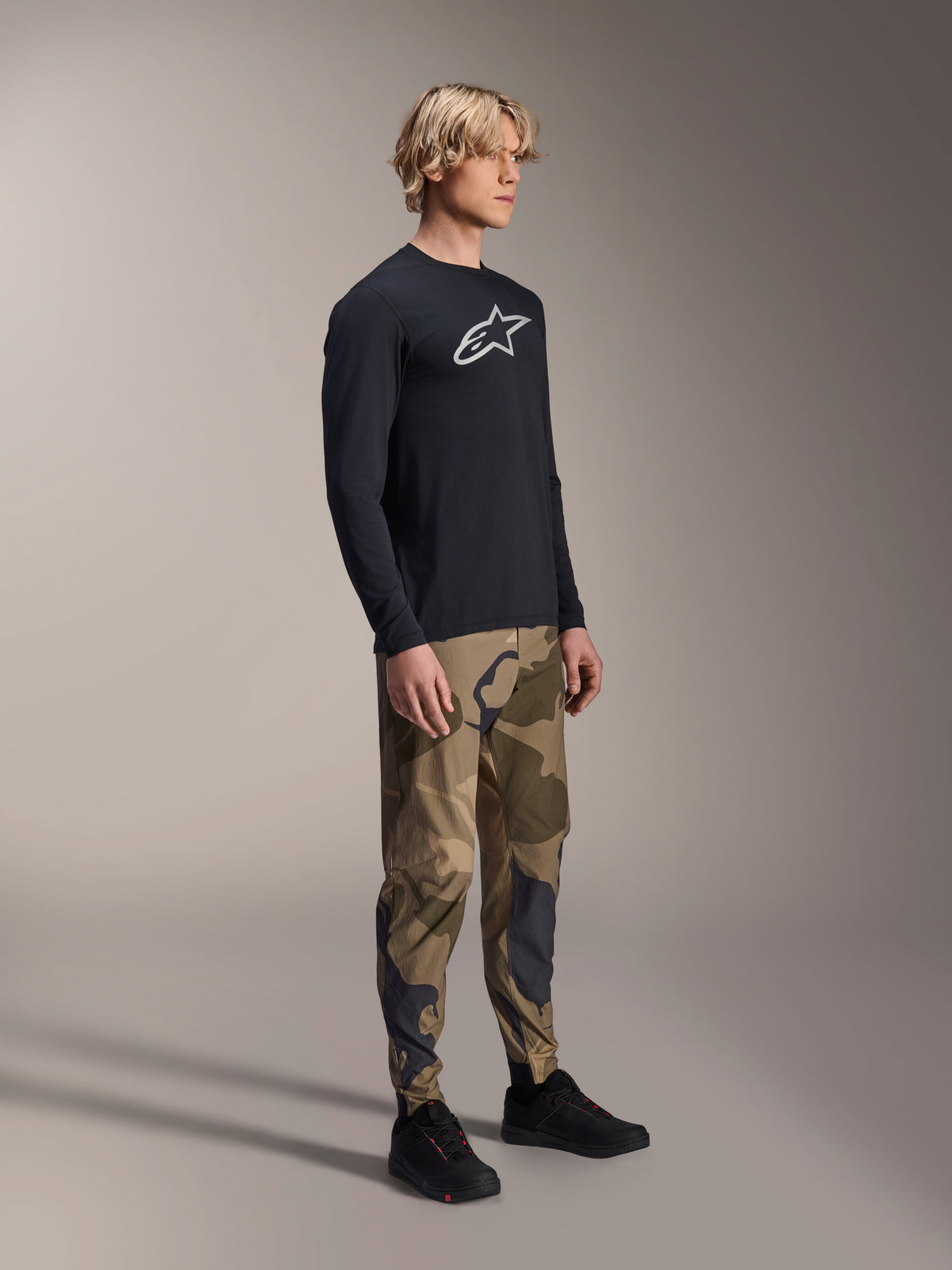 Alpinestars A-Dura Astar Long Sleeve Jersey in Black, worn by a model with camouflage pants and black sneakers, featuring the white Astars chest logo, casual performance MTB apparel for trail riding.