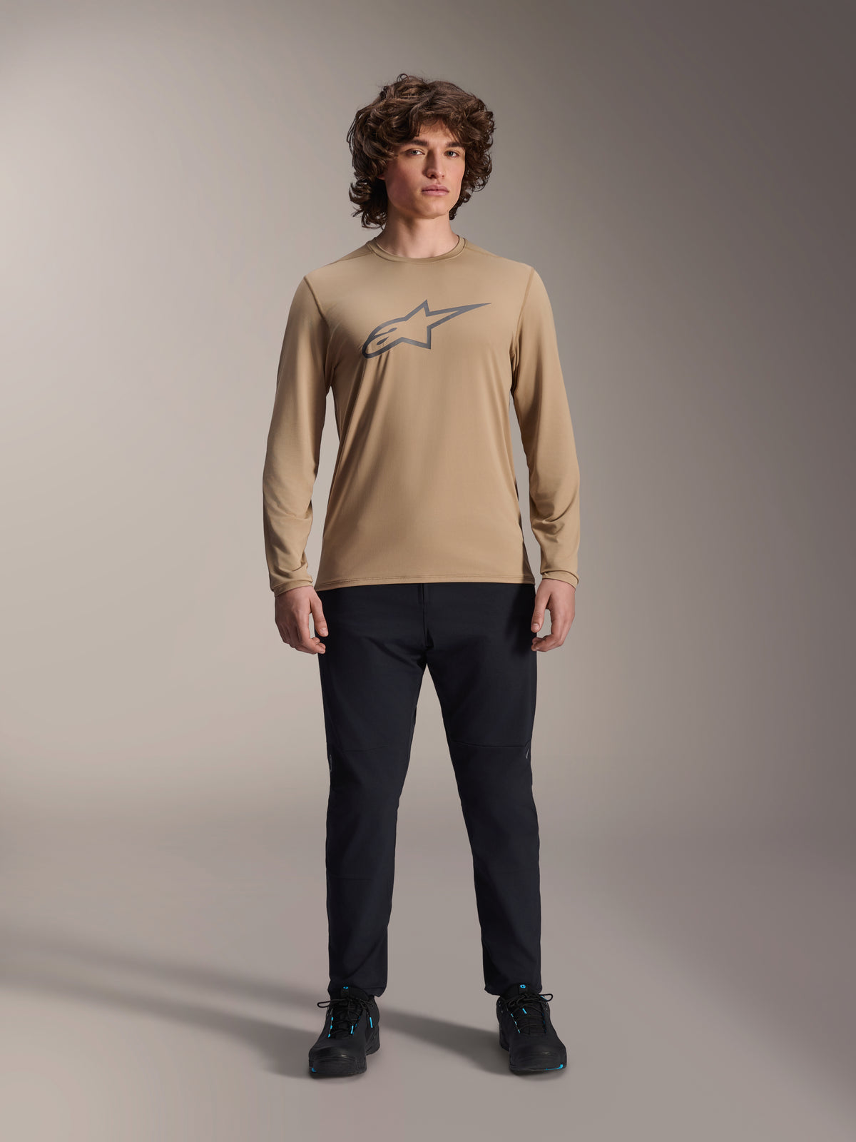 Alpinestars A-Dura Astar långärmad tröja, ljusbrun med grå Astars-logotyp på bröstet, bärs av en modell med svarta tekniska byxor och skor, avslappnad prestationsutrustning för mountainbike