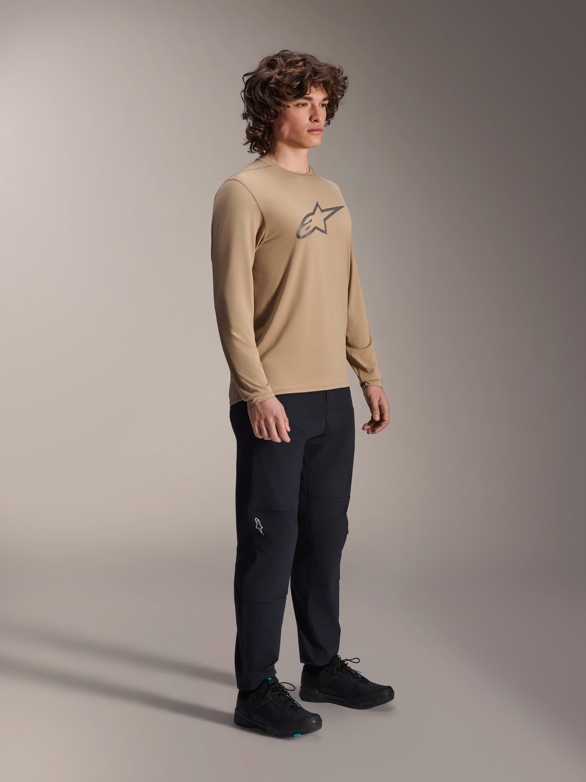 Alpinestars A-Dura Astar långärmad tröja, ljusbrun, bärs av en modell med svarta byxor och skor, med en grå Astars-logotyp på bröstet, avslappnad prestandautrustning för mountainbike