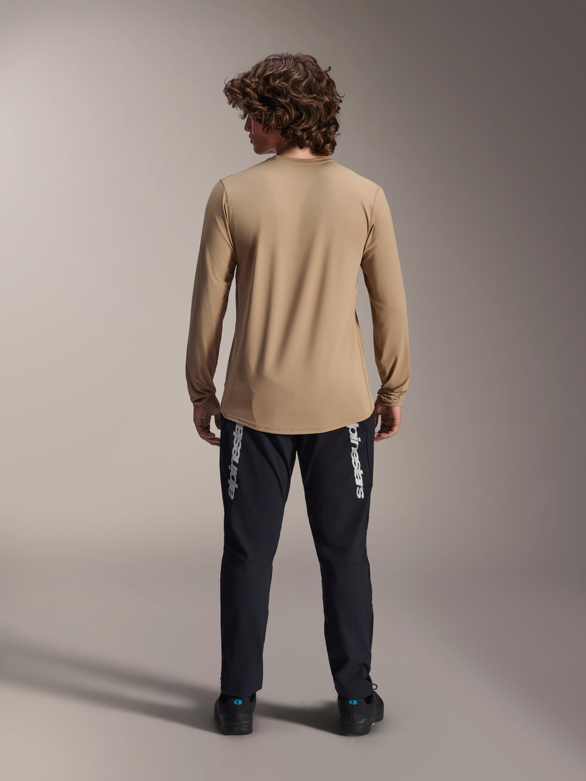 Alpinestars A-Dura Astar långärmad tröja, ljusbrun, vy bakifrån som visar den förlängda ryggen, bärs med svarta byxor med Alpinestars-logotyp, tekniska mountainbikekläder