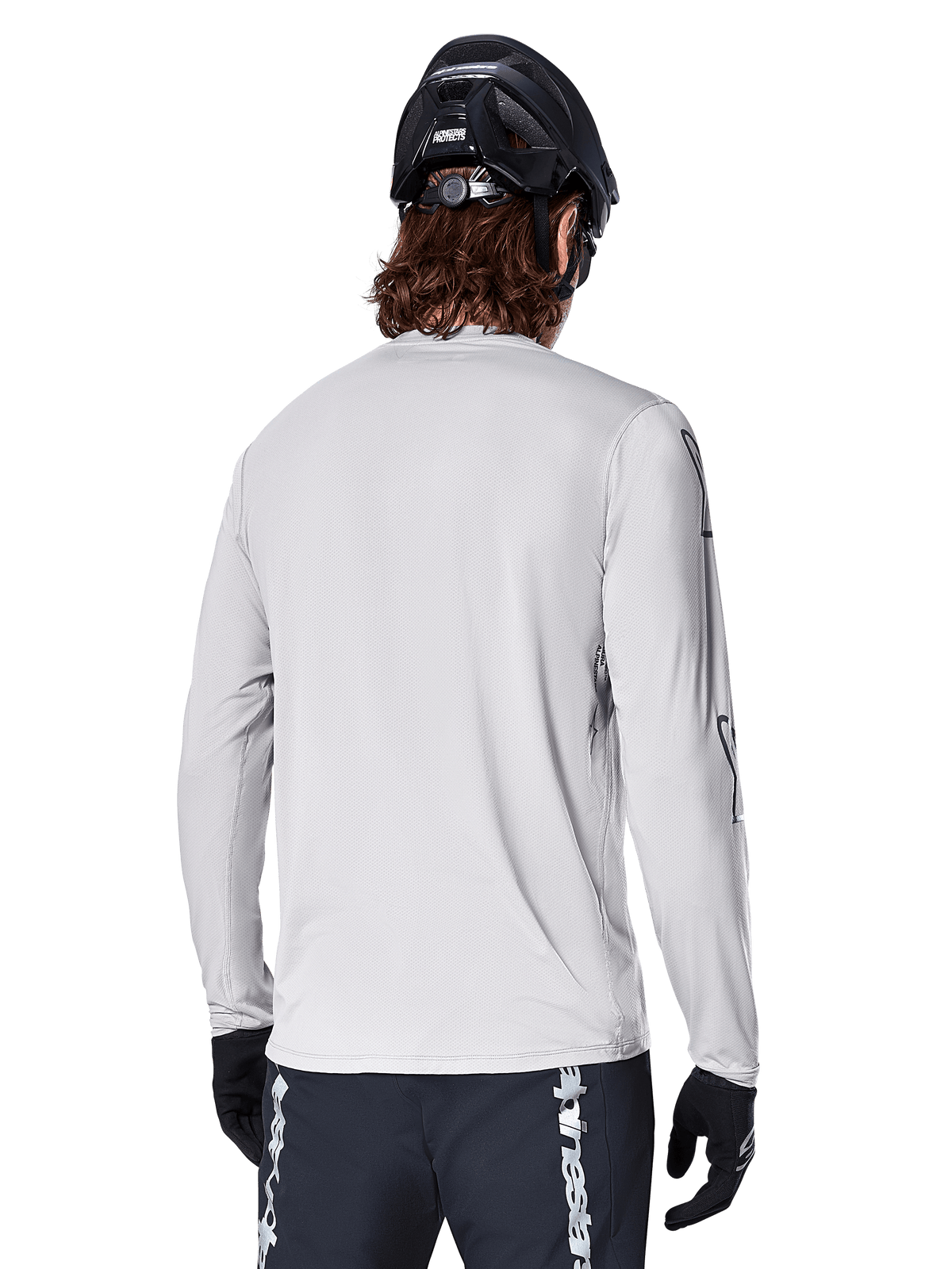 Alpinestars A-Dura Switch långärmad tröja, ljusgrå, bakifrån sett på en mountainbikecyklist med svart MTB-hjälm och handskar, har ett förlängt bakstycke för extra skydd och strukturerat jacquardtyg, teknisk cykelutrustning.