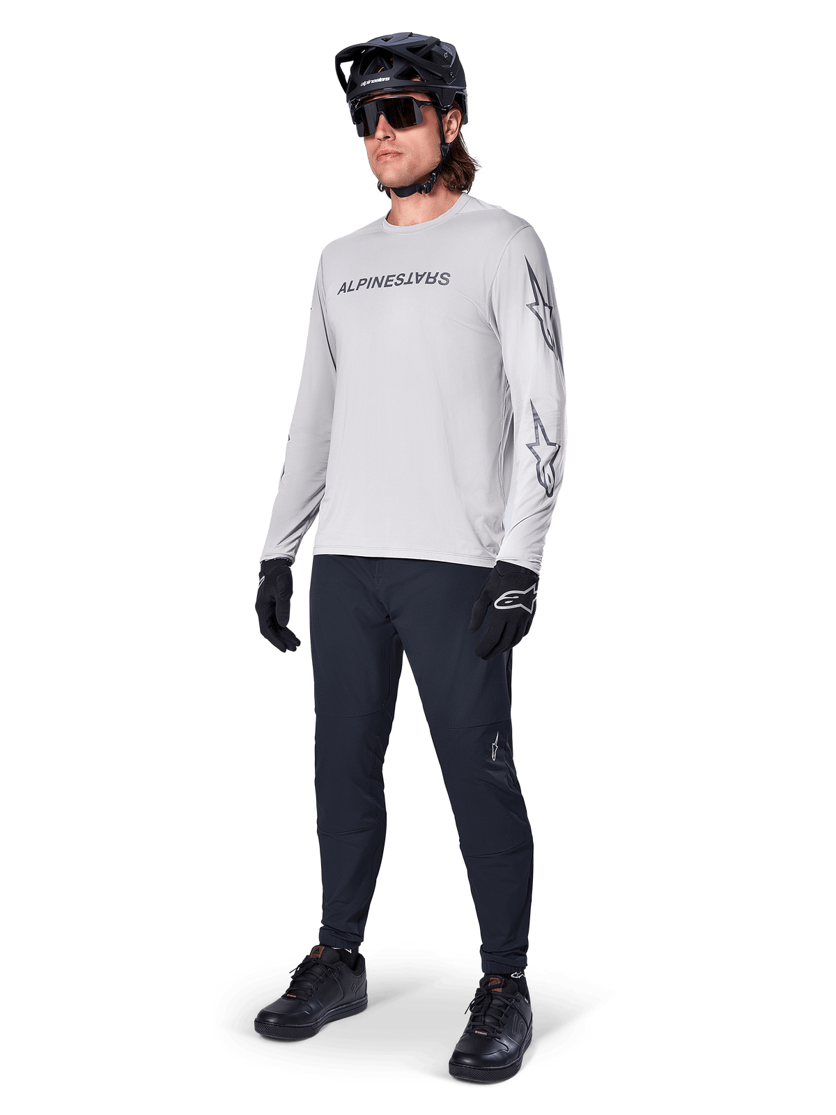 Alpinestars A-Dura Switch långärmad tröja, ljusgrå, bärs av manlig cyklist med svarta mountainbikebyxor och hjälm, med svart logotyp på bröstet och stjärngrafik på ärmarna, högpresterande cykelutrustning för terrängkörning