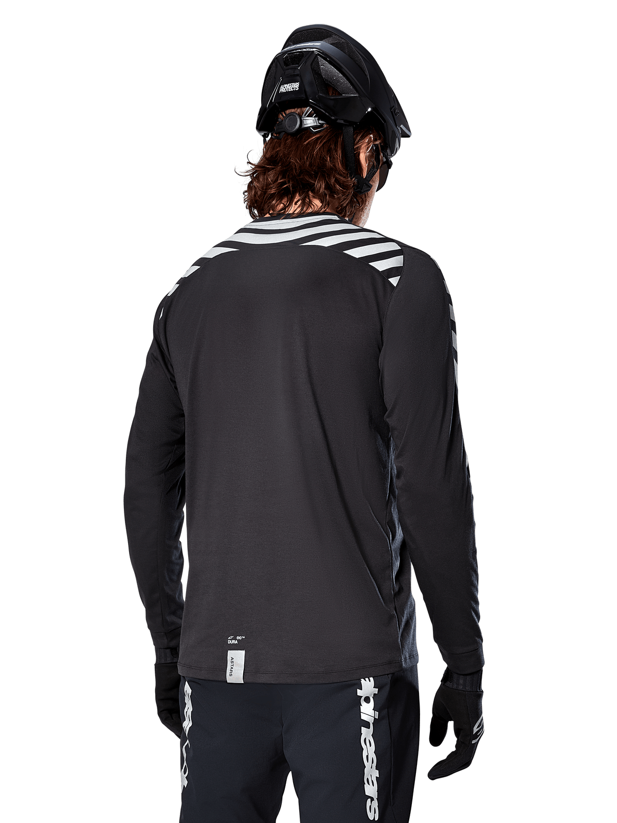Alpinestars A-Dura Dri Oscar långärmad tröja, Black Reflective, vy bakifrån som visar reflekterande diagonala ränder på axlarna och ett förlängt ryggslut, buren med svart MTB-hjälm och handskar, teknisk utrustning för mountainbike
