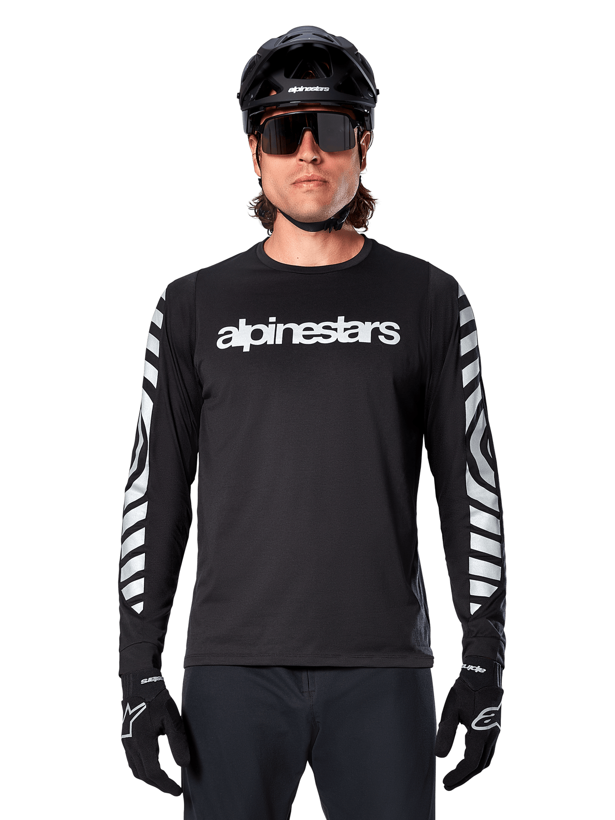 Manlig cyklist i Alpinestars A-Dura Dri Oscar långärmad tröja, Black Reflective, med vit Alpinestars-text på bröstet och reflekterande silvergrafik på ärmarna, kombinerat med svart MTB-hjälm, solglasögon och svarta handskar, teknisk cykelutrustning