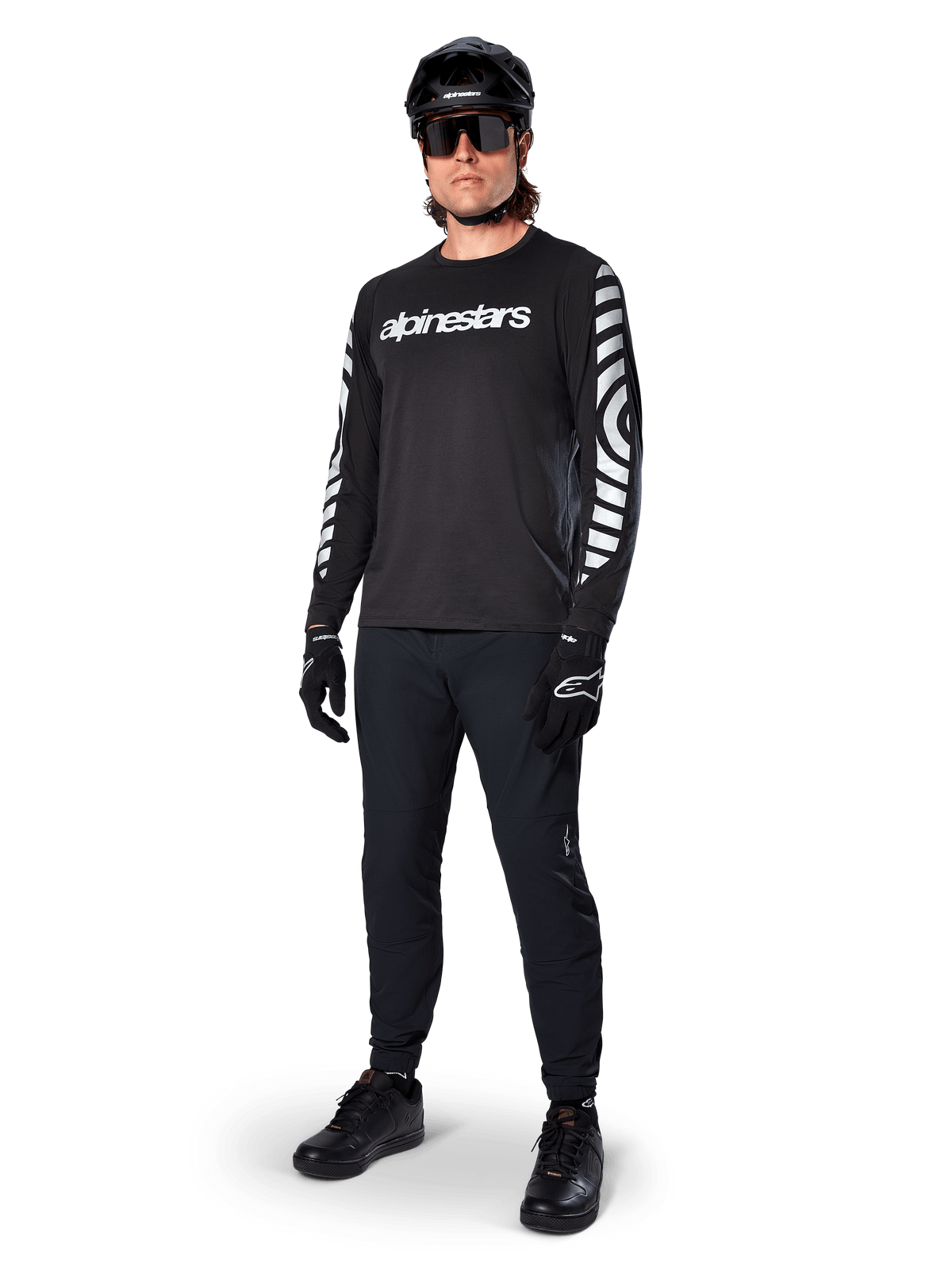 Alpinestars A-Dura Dri Oscar långärmad tröja, Black Reflective, med en tydlig vit Alpinestars-textlogotyp på bröstet och reflekterande grafiska mönster på ärmarna, bärs med svarta mountainbikebyxor, hjälm och handskar, tekniska MTB-kläder