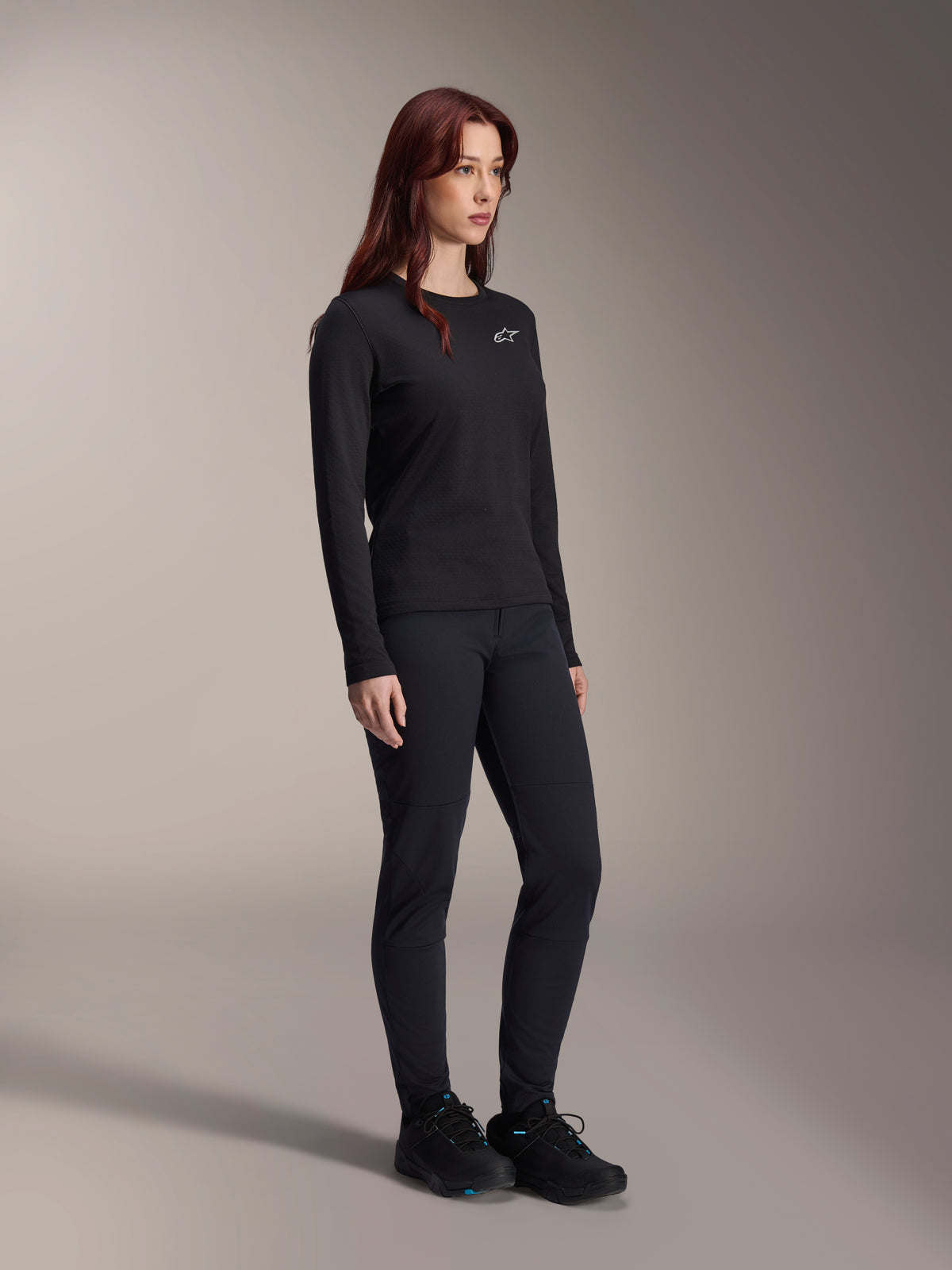 Kvinna som bär Alpinestars Stella A-Dura Thermal Jersey, långärmad svart MTB-tröja med vit logotyp på bröstet, stylad med matchande svarta byxor och sportskor, studioporträtt för funktionella cykelkläder