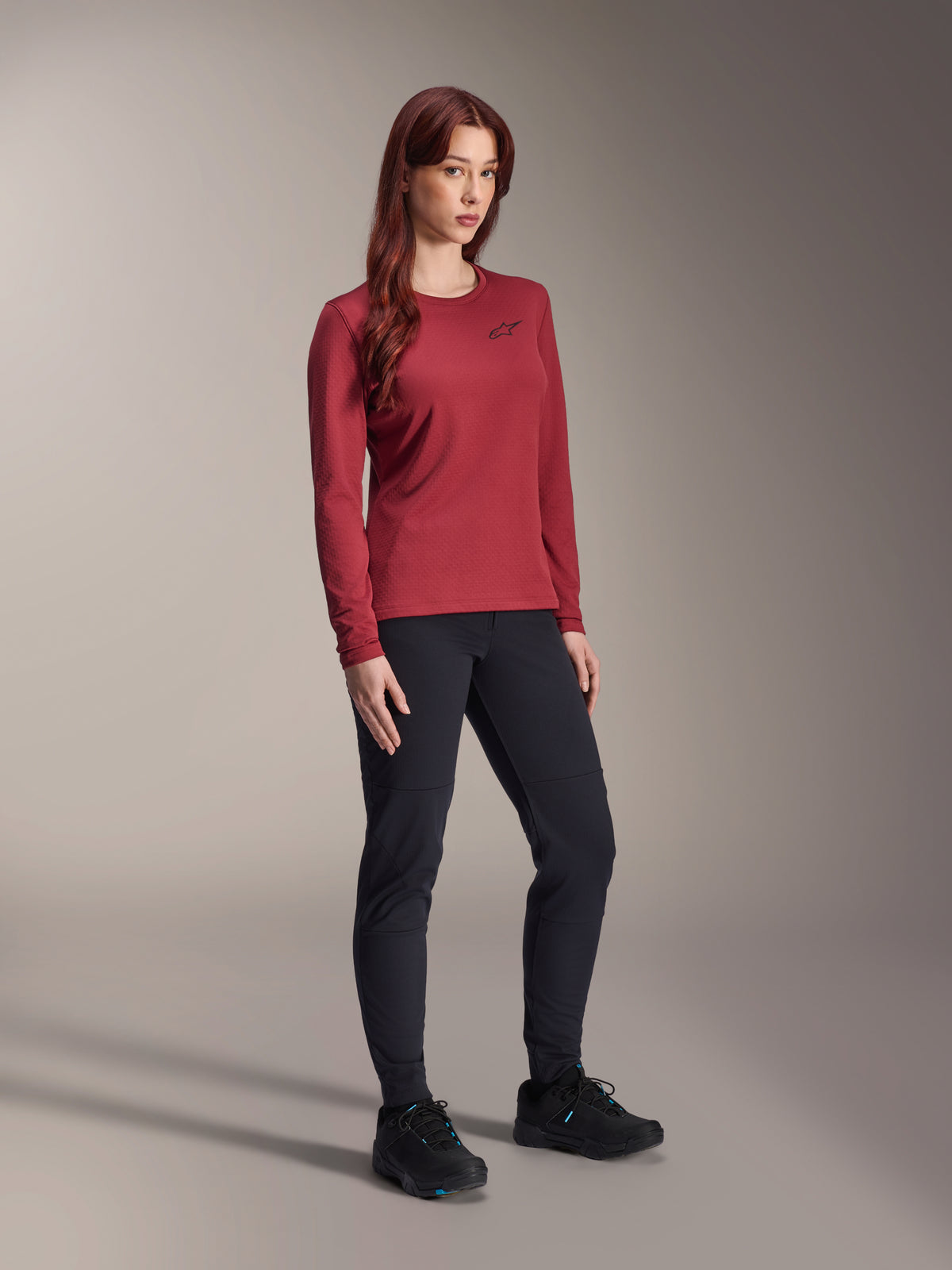 Kvinna som bär svarta Alpinestars Stella A-Dura Thermal-byxor och en vinröd långärmad Stella A-Dura Thermal-tröja, tekniska mountainbikekläder för cykling i kallt väder, slim fit-design med slitstarkt material, studiobakgrund