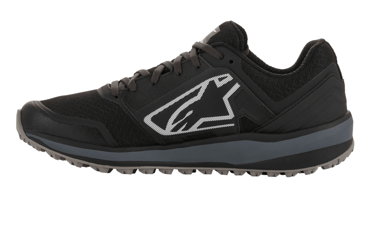 Alpinestars Meta Trail-skor, prestationslöparsko, svarta och mörkgrå, sidoprofil med stor vit Astars-logotyp, mörkgrå mellansula, räfflad oljebeständig gummisula, designad för komfort i paddocken och för motorsportsteam