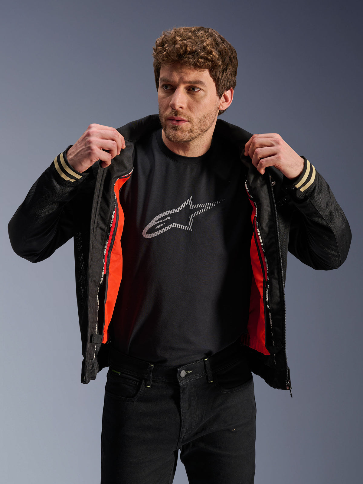 Man som bär Alpinestars Ignite Air-jacka i svart, med meshkonstruktion och ett klart orange avtagbart vindskyddsfoder, kombinerat med en svart t-shirt med Astars-logotyp och svarta byxor, urban samtida motorcykelstil