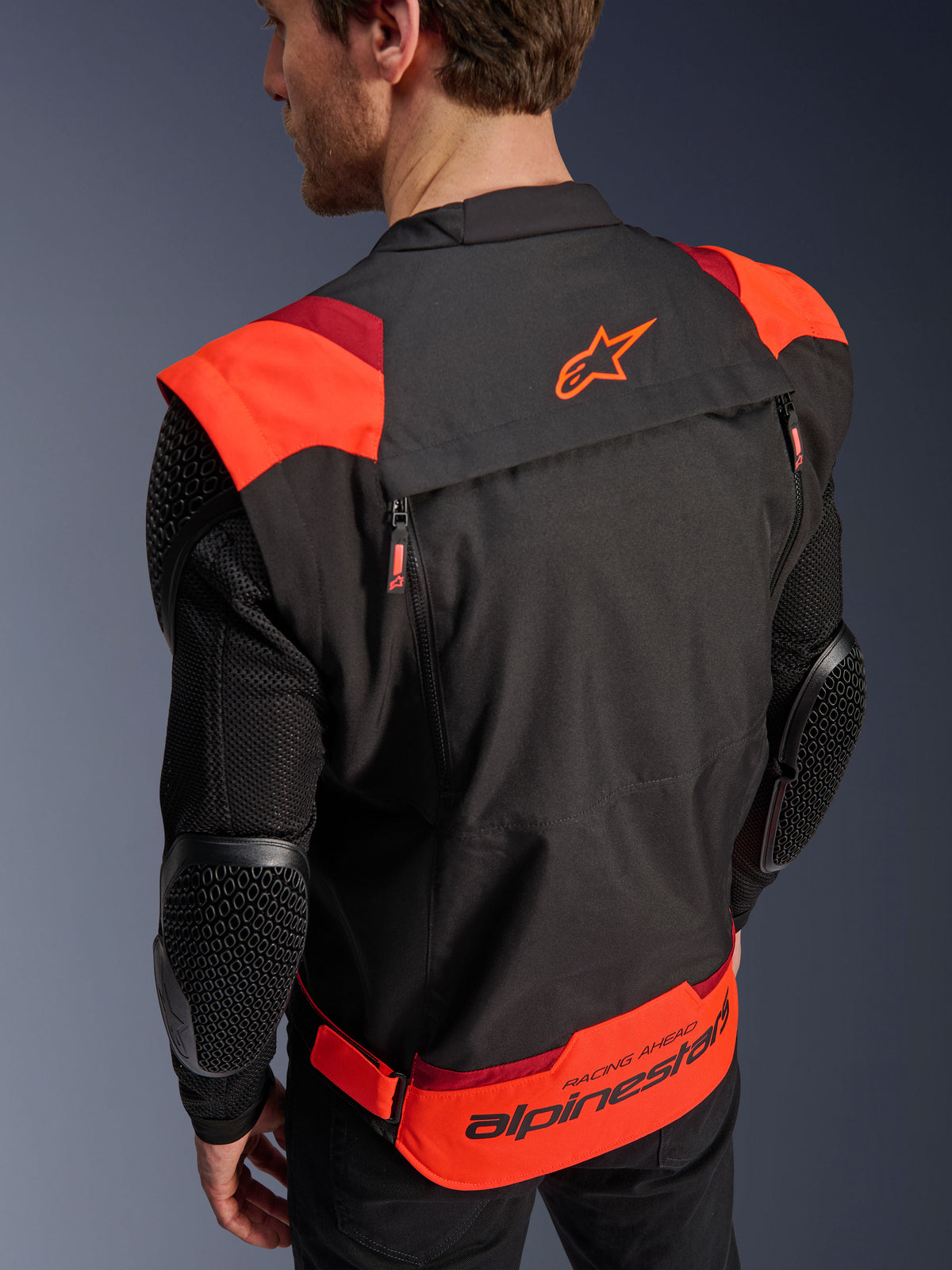 Alpinestars T-Stunt Air Jacket, Black Red Fluo and Dark Red, rear view on a model, showing mesh sleeves with honeycomb-textured elbow protectors, Alpinestars star logo on the upper back, and bold lower back branding with 'RACING AHEAD' text, modular sport-cut design for motorcycle riding.