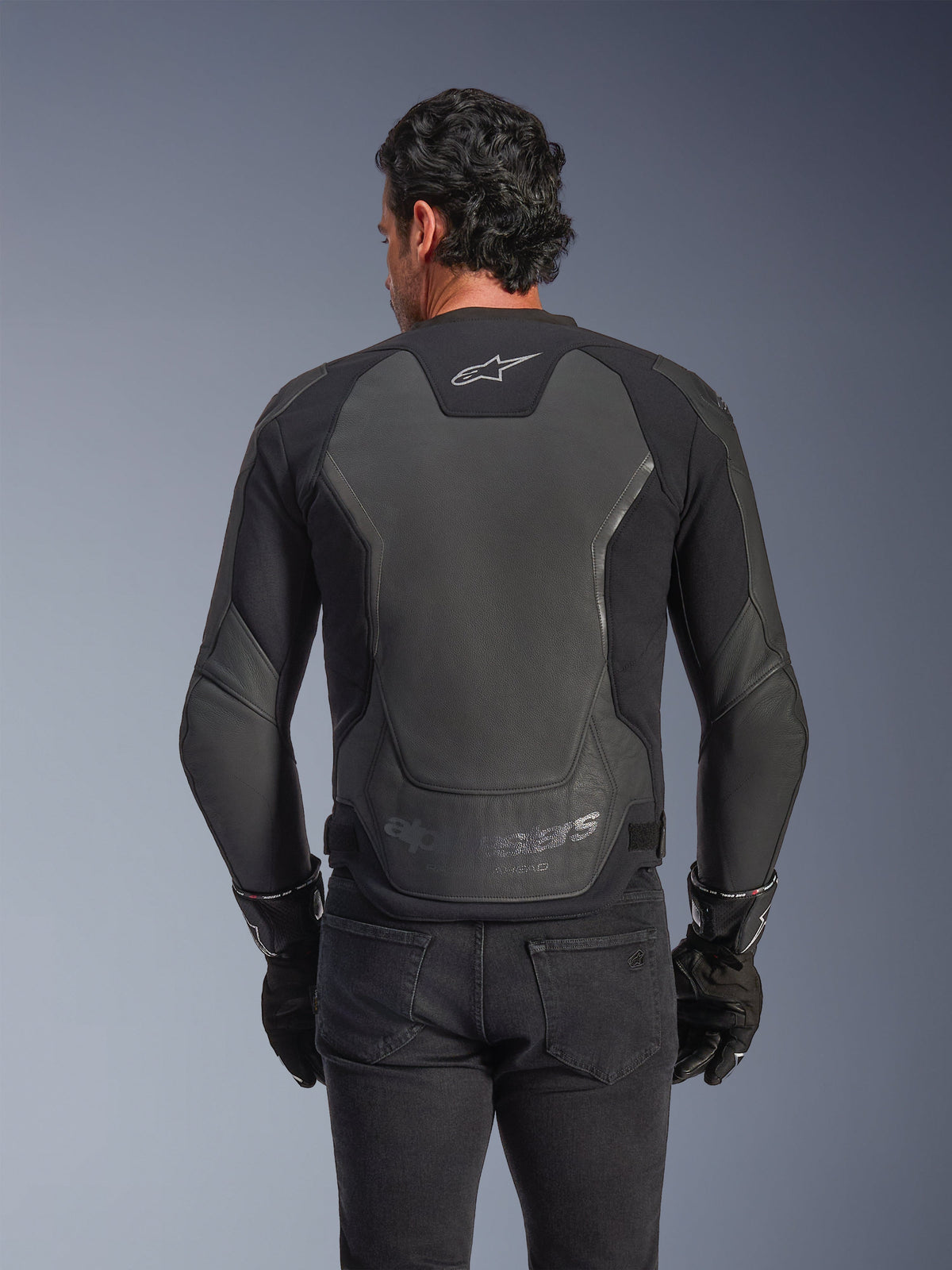 Alpinestars GP Force V2 läderjacka, svart, vy bakifrån av en man som bär jackan med svarta handskar och jeans, med stora skyddspaneler i läder på ryggen, Alpinestars-logotyp på övre delen av ryggen och präglad logotyp vid nedre delen av midjan, sportinspirerad motorcykelutrustning