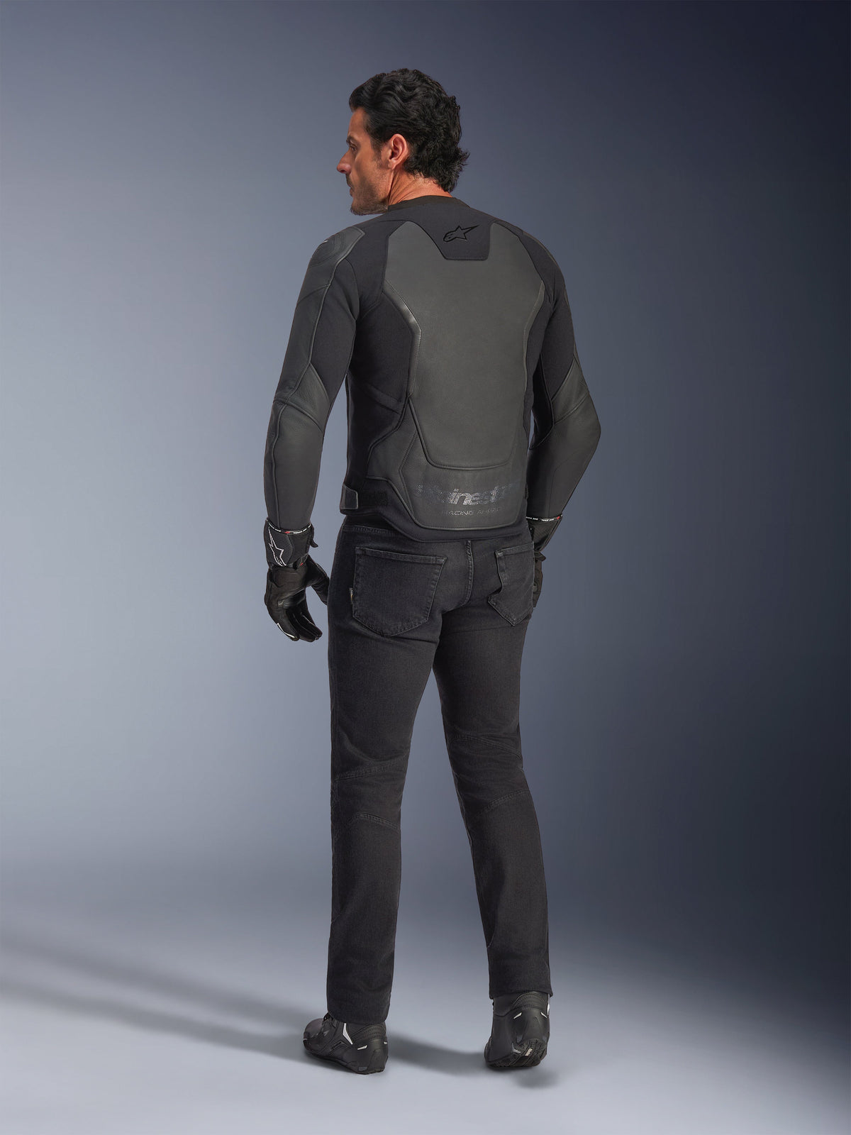 Alpinestars GP Force V2 läderjacka i Black Black, sedd bakifrån på en modell, med flerpanelkonstruktion i läder och sportpassform, kombinerad med svarta körbyxor, handskar och stövlar för en komplett racing-inspirerad look.