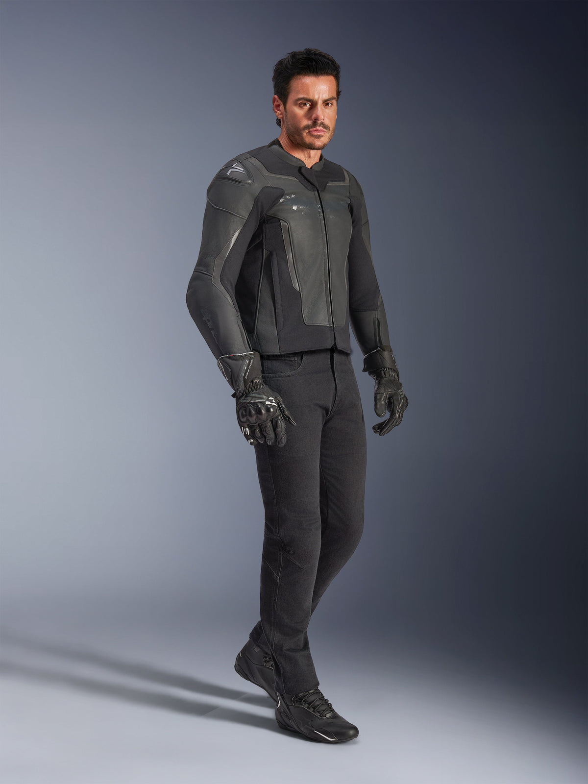 Man i Alpinestars GP Force V2 läderjacka i svart, tillsammans med svarta byxor och motorcykelhandskar, helkroppsbild i studiomiljö, smidig konstruktion i nötläder med integrerat axel- och armbågsskydd, professionell sportutrustning för motorcykel