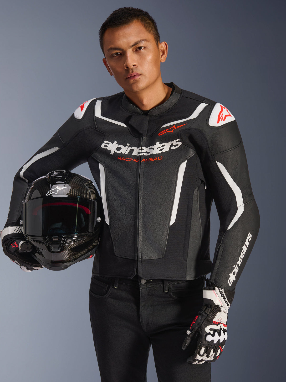 Alpinestars GP Force V2 läderjacka, svart och vit med röda detaljer, bärs av en modell som håller en motorcykelhjälm i kolfiber och bär racinghandskar, med vita kontrastpaneler och Alpinestars logotyp.