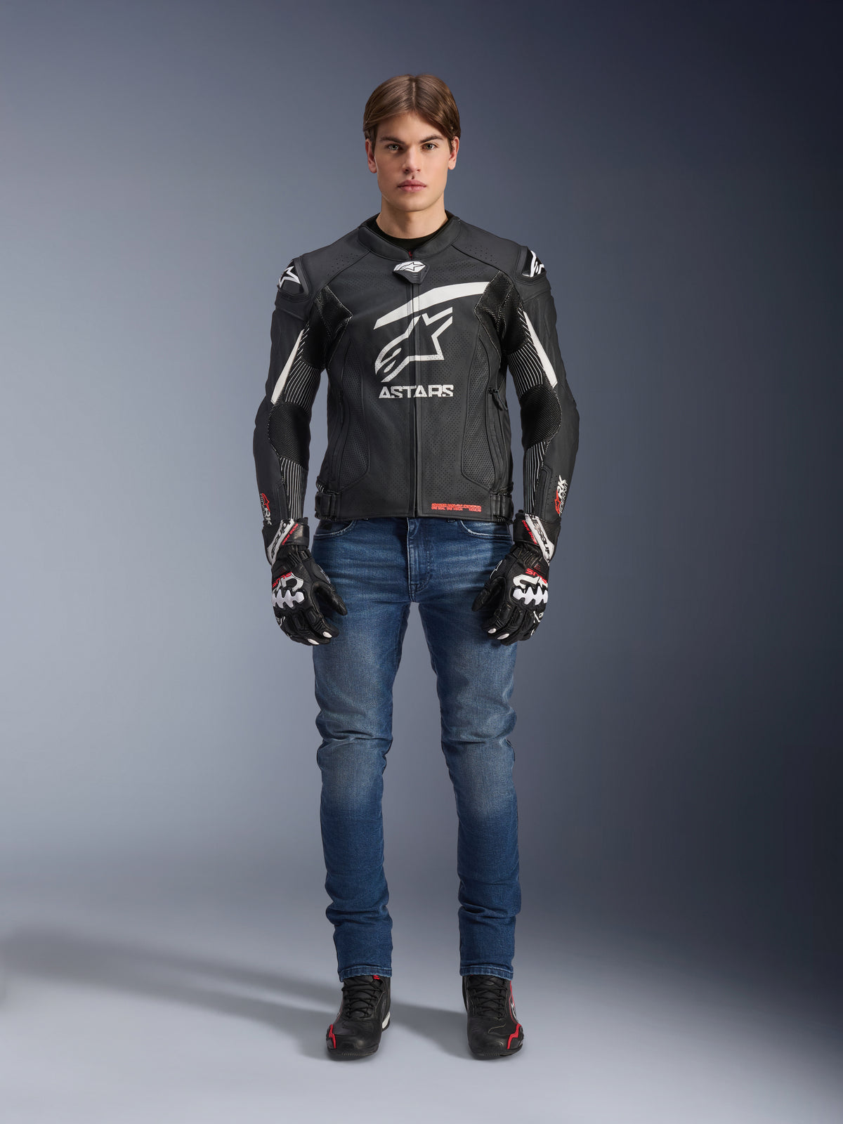 Manlig modell bär Alpinestars GP Plus R V4 Rideknit läderjacka i svart och vitt, stylad med blå MC-jeans, svarta motorcykelhandskar och svarta MC-skor, med en stor vit Astars-logotyp på bröstet, perforerade läderpaneler och vita racingränder på ärmarna, stående helkroppsbild