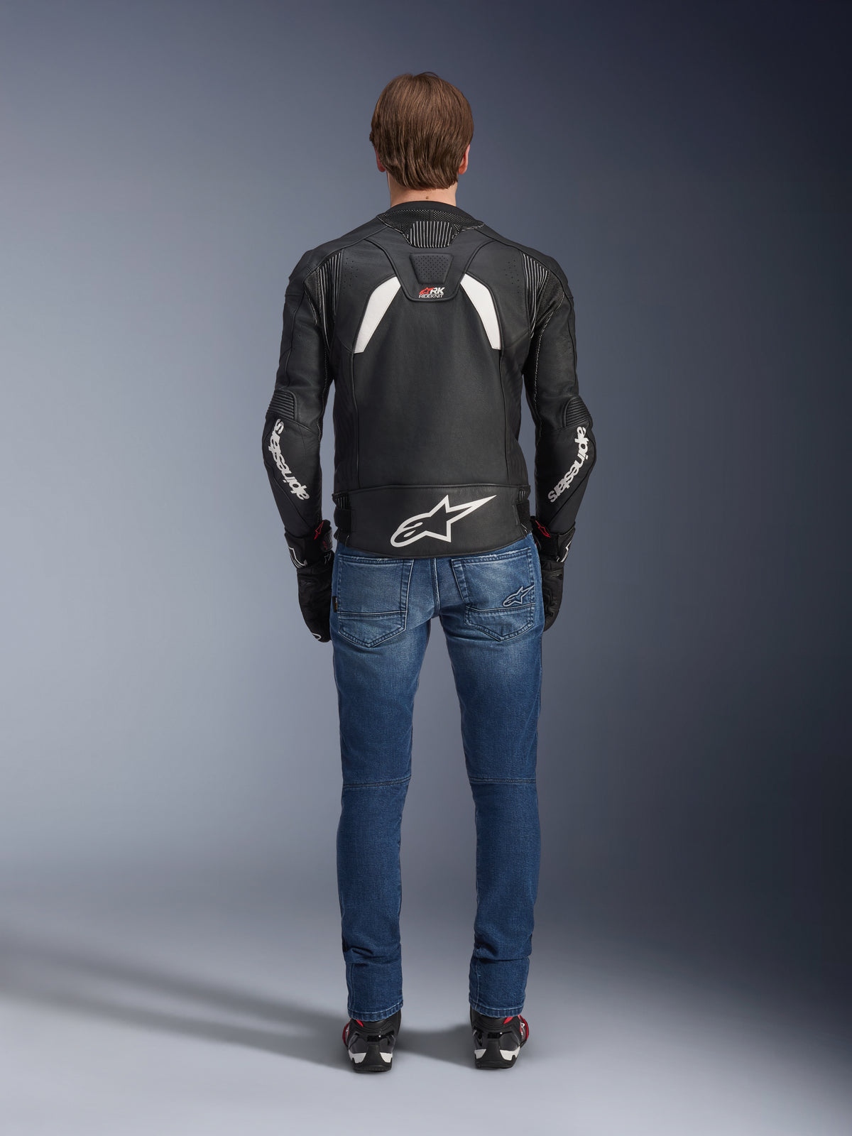 Alpinestars GP Plus R V4 Rideknit läderjacka sedd bakifrån, svart och vit, med en stor vit logotyp på nedre delen av ryggen och logotyper på ärmarna, bärs med blå mc-jeans och svarta stövlar, vilket framhäver den tekniska konstruktionen och sportiga passformen.