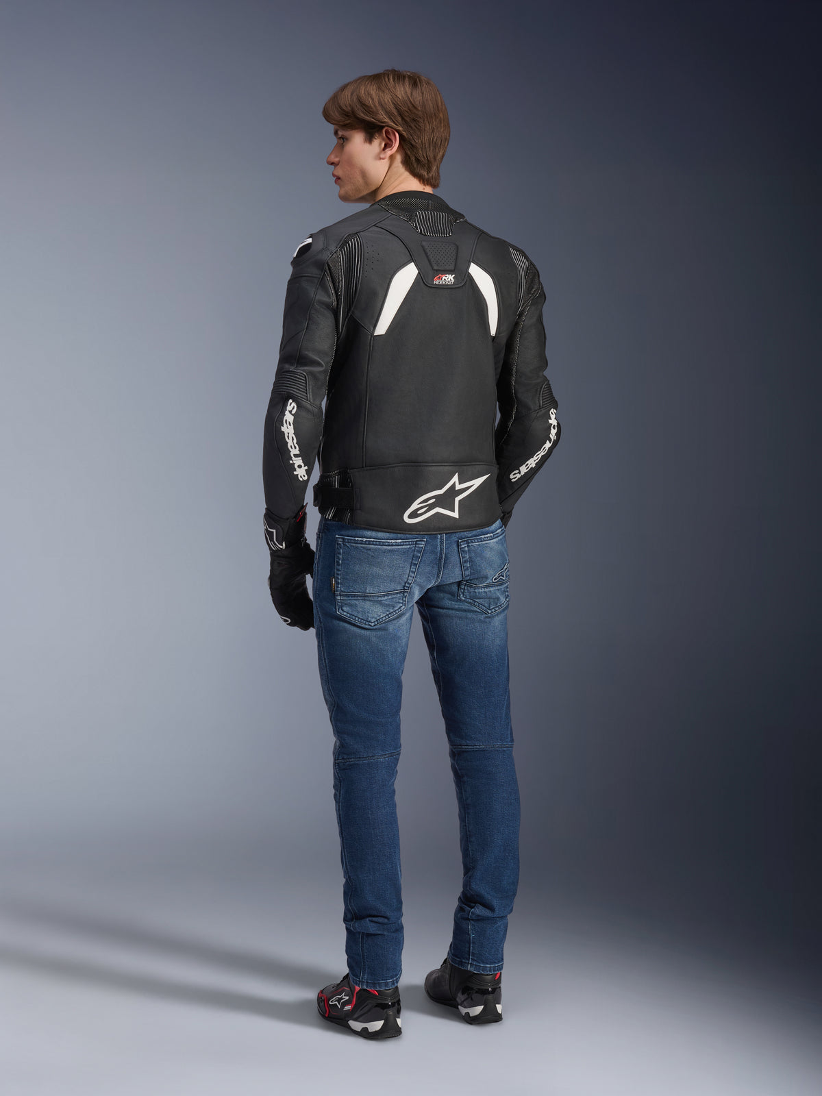 Alpinestars GP Plus R V4 Rideknit-läderjacka i svart och vitt, sedd bakifrån på en modell, med vit Alpinestars-branding på ärmarna och en stor stjärnlogotyp på nedre delen av ryggen, bärs med blå jeans och svarta mc-skor, teknisk motorcykelutrustning.