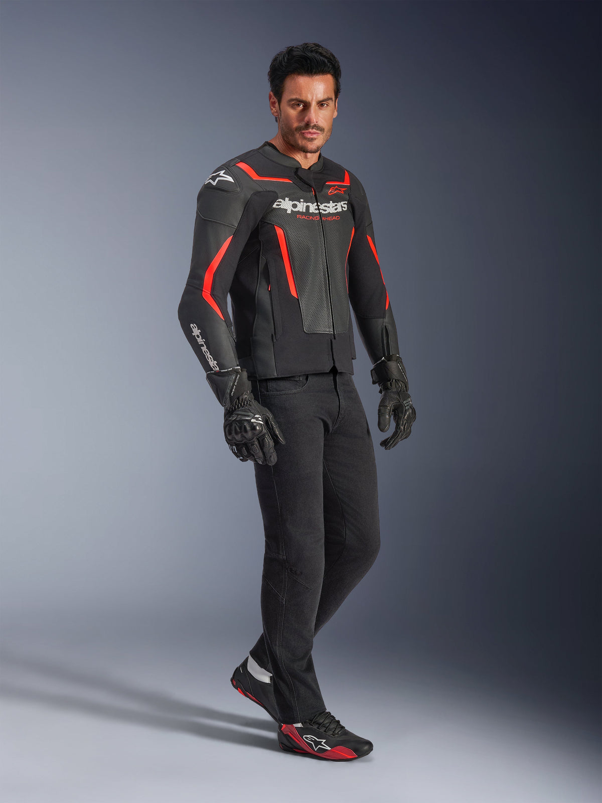 Modell som bär Alpinestars GP Force V2 Air läderjacka i svart och neonrött, matchad med svarta körbyxor, handskar och matchande sneakers, med perforerade läderpaneler och framträdande vit Alpinestars-logotyp, sportmotorcykelutrustning