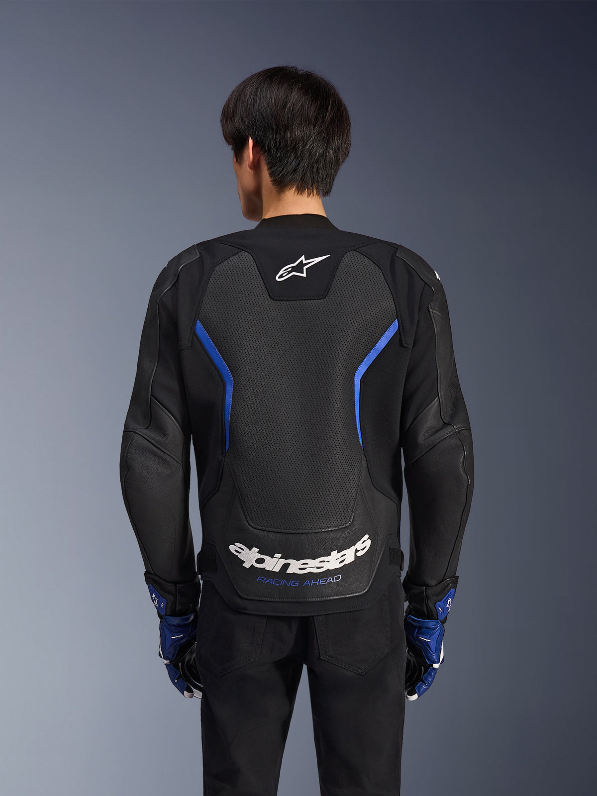 Alpinestars GP Force V2 Airflow läderjacka, Black Metallic Blue, vy bakifrån, med en stor perforerad ryggpanel för ventilation, blå detaljer och vit Alpinestars-logotyp, bärs med matchande blå handskar och svarta byxor för sportkörning.