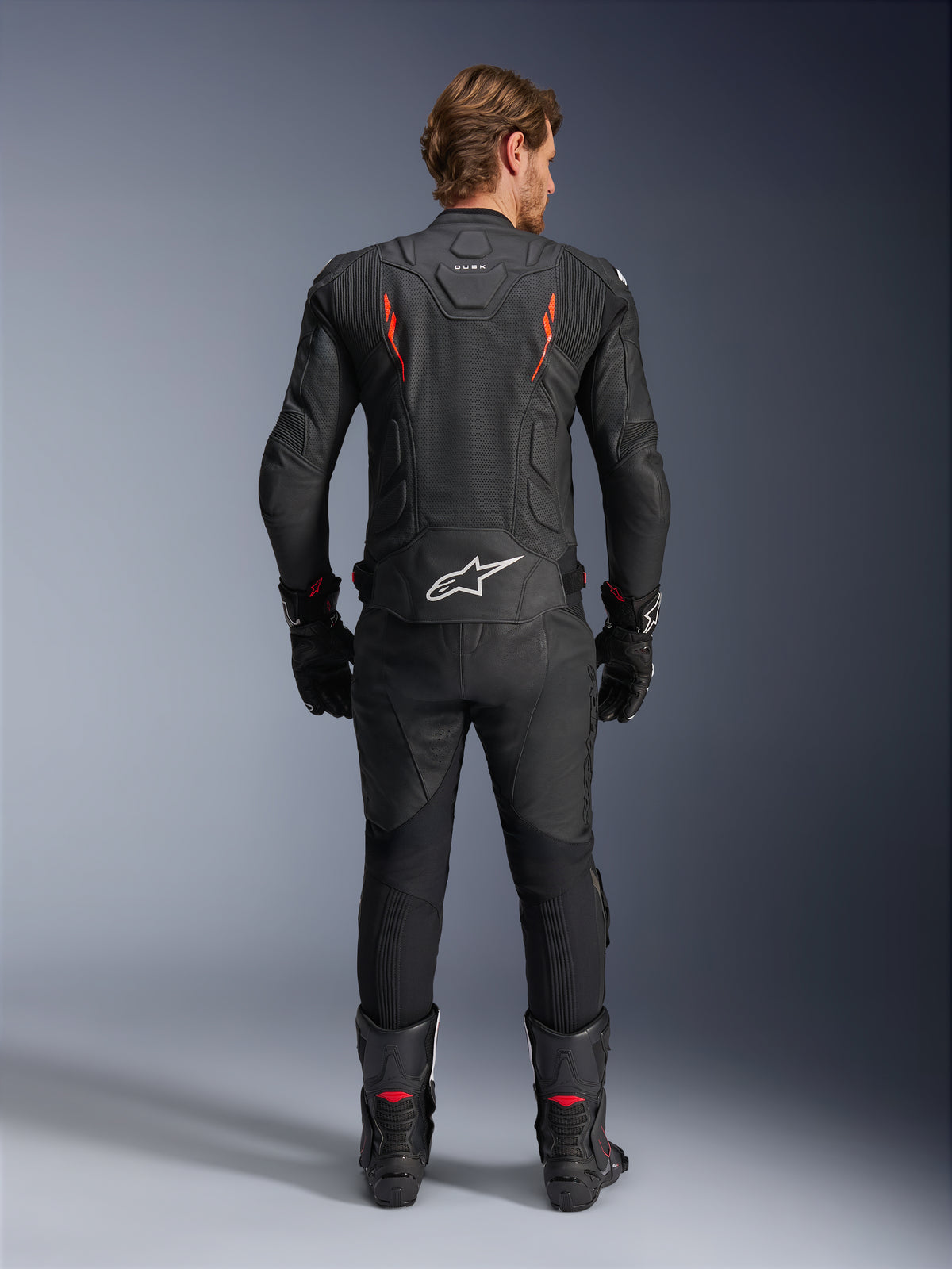 Alpinestars Dusk Airflow läderjacka, svart och röd fluo, baksida med omfattande perforeringar och stor vit logotyp på nedre delen av ryggen, bärs med matchande racingutrustning i läder inklusive byxor, stövlar och handskar