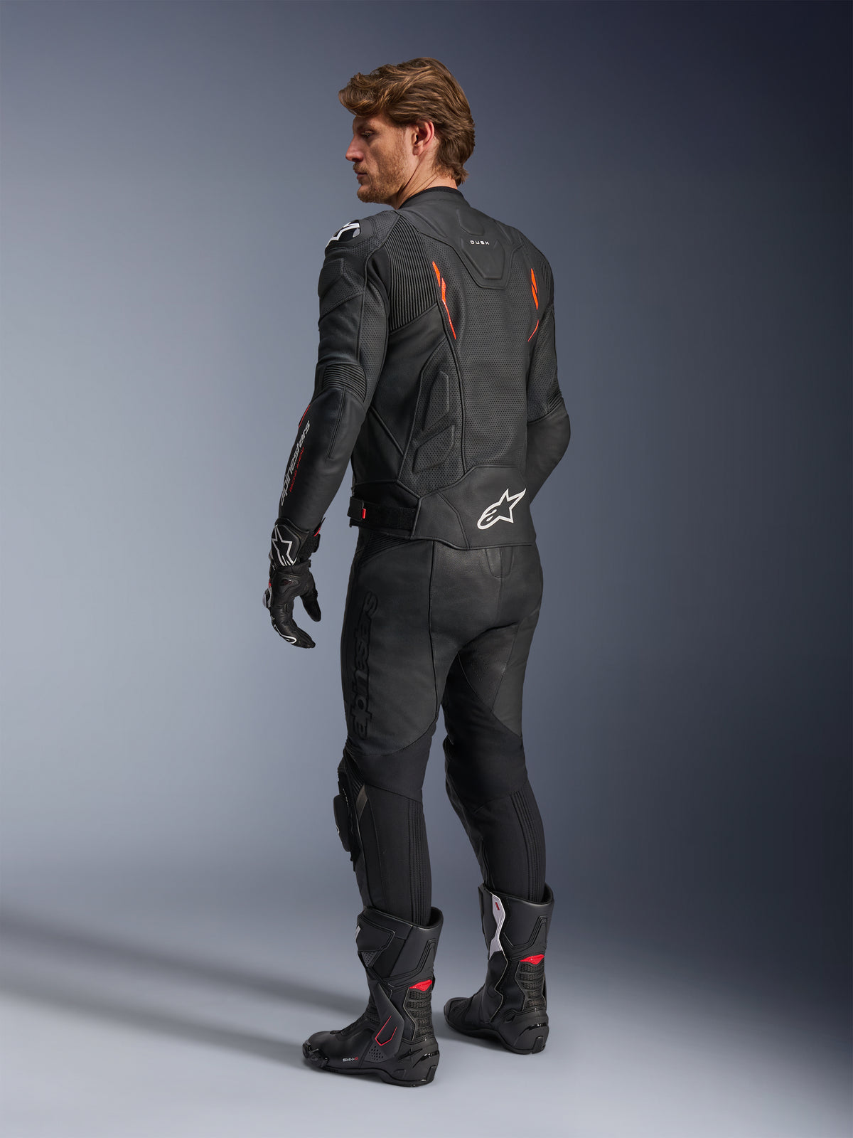 Alpinestars Dusk Airflow läderjacka, svart med Red Fluo-detaljer, ryggvy av en förare i full motorcykelutrustning i läder inklusive matchande svarta byxor, handskar och racingstövlar, med perforerat läder och vita Alpinestars-logotyper.