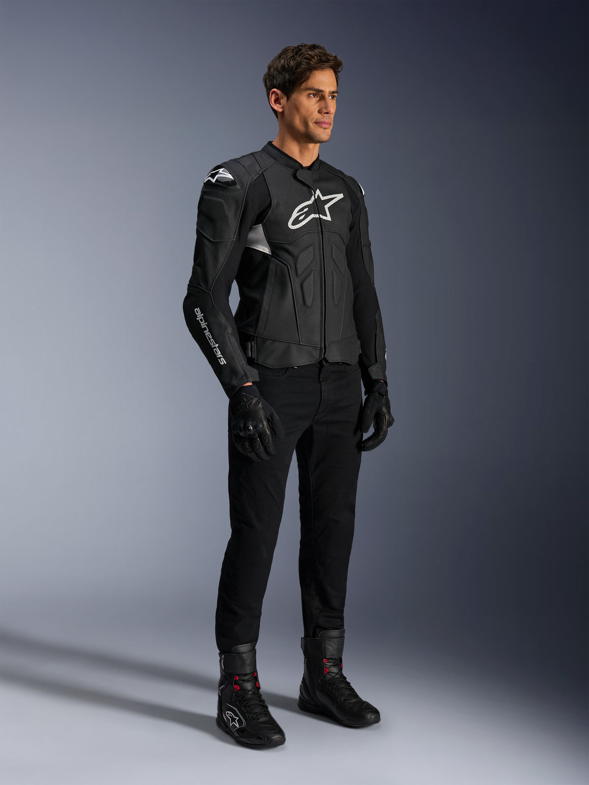 Alpinestars Dusk Airflow läderjacka, svart och vit, modellerad med svarta motorcykelbyxor, handskar och stövlar, med perforerade läderpaneler och vita logotyper, sportig road racing-stil.