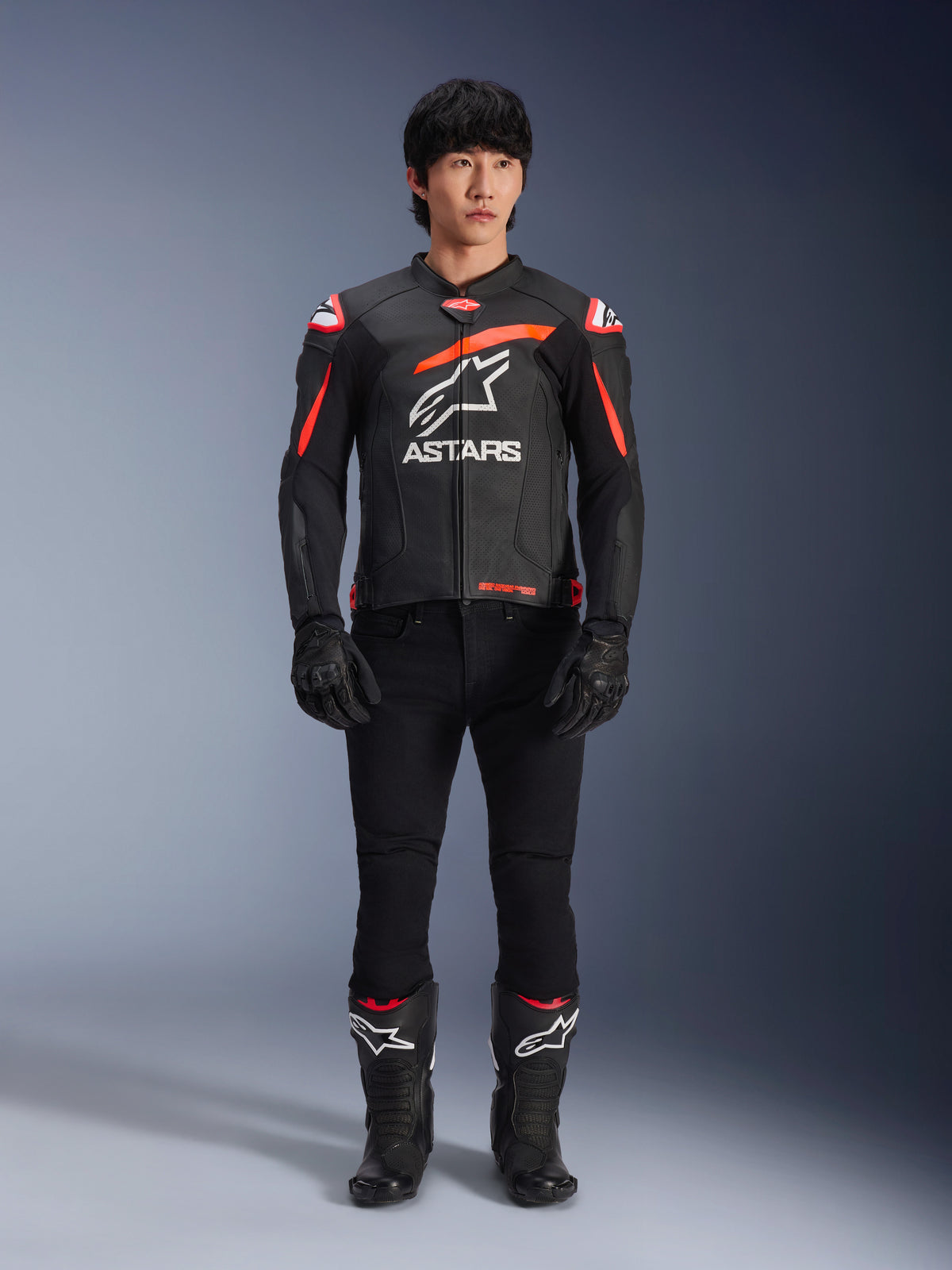 Manlig modell i Alpinestars GP Plus R V4 Airflow skinnjacka, svart med detaljer i Red Fluo och vitt, stor vit Astars-logotyp på bröstet, kombinerad med svarta tekniska byxor, handskar och stövlar, prestandautrustning för motorcykel i studiomiljö