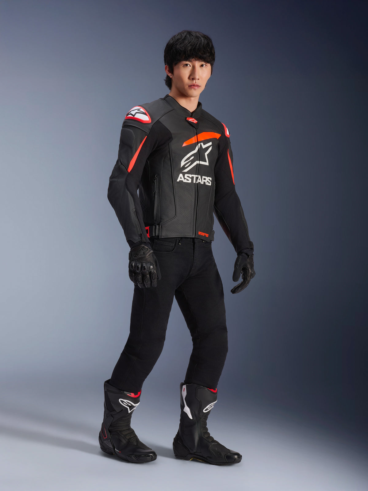 Manlig förare iförd Alpinestars GP Plus R V4 Airflow läderjacka, svart med Red Fluo- och vita detaljer, med perforerat läder och ASTARS-logotyp på bröstet, bärs med svarta mc-byxor och svarta motorcykelstövlar, sportprestandautrustning