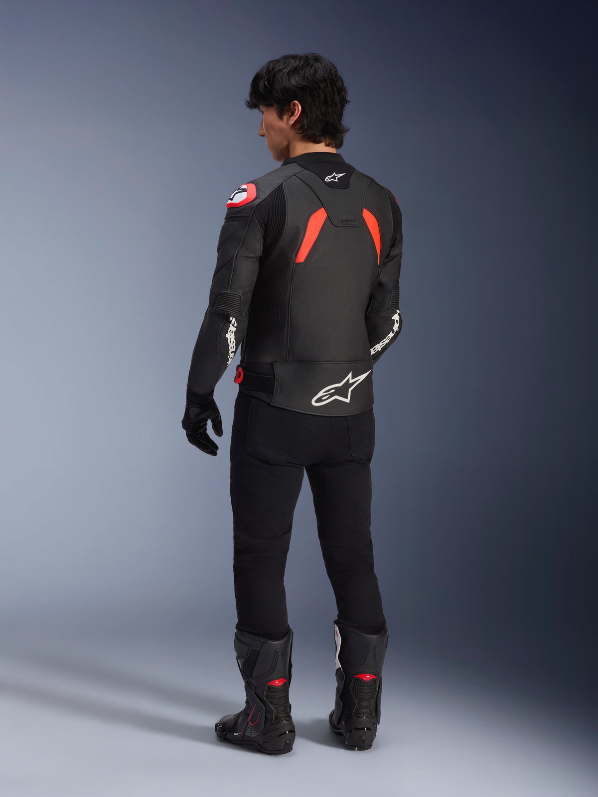 Bakifrån vy av en modell som bär Alpinestars GP Plus R V4 Airflow läderjacka, svart med röd fluo och vita detaljer, kombinerat med svarta byxor och svarta motorcykelstövlar, framträdande vita logotyper på ryggen och ärmarna, sportig läderkonstruktion.