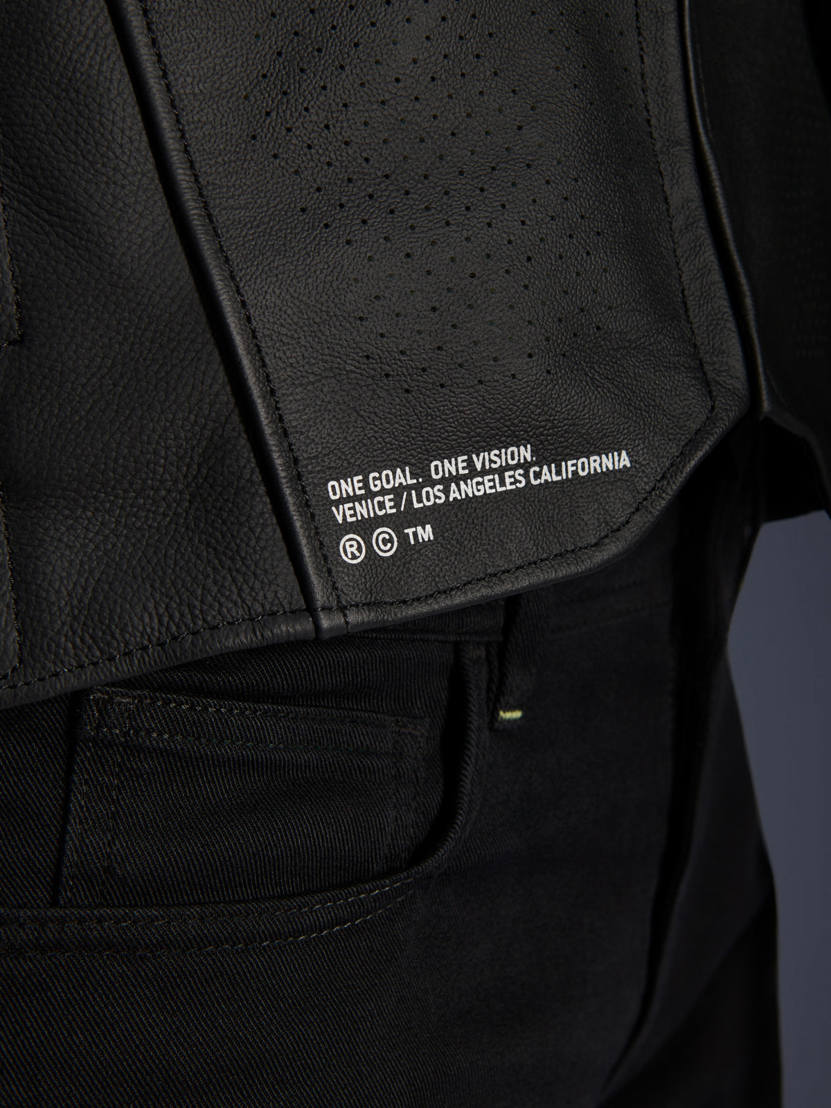 Närbild av Alpinestars Atem V5 läderjacka i svart, med den nedre ryggpanelen, 'ONE GOAL. ONE VISION. VENICE / LOS ANGELES CALIFORNIA'-tryck och perforerad läderstruktur.