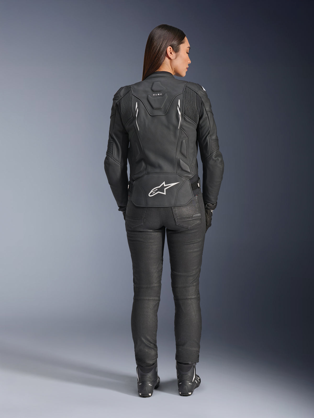 Alpinestars Stella Dusk läderjacka för dam, svart, sedd bakifrån med vit Alpinestars-logotyp på nedre delen av ryggen och 'DUSK'-märkning på övre delen av ryggen, buren med svarta MC-byxor och stövlar, framhäver den konturerade dampassformen och den skyddande läderkonstruktionen.