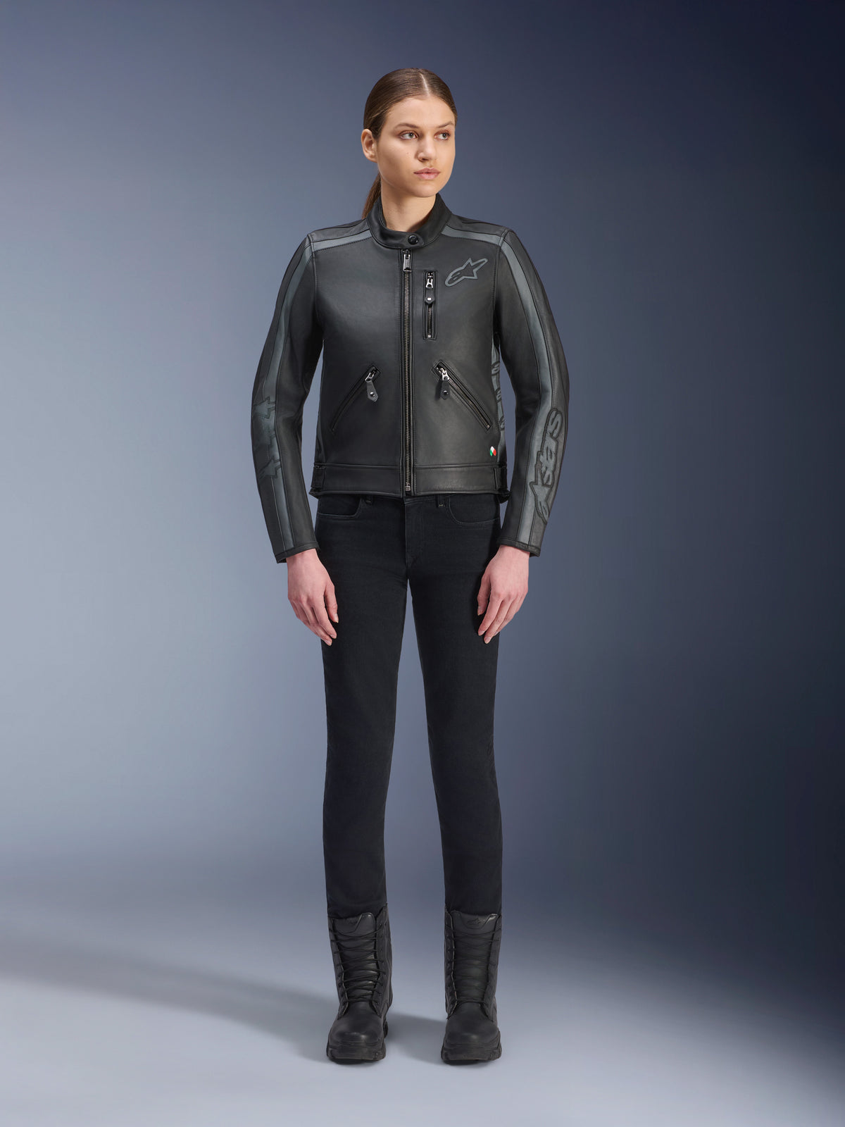 Alpinestars Stella Dyno läderjacka, motorcykeljacka för dam, Black Tar Gray Dark Gray, studiobild i helfigur av en modell som bär jackan med svarta slim-fit-byxor och svarta stövlar, tillverkad i nötläder med grå detaljer och diskret Alpinestars-logotyp på bröstet och ärmarna.