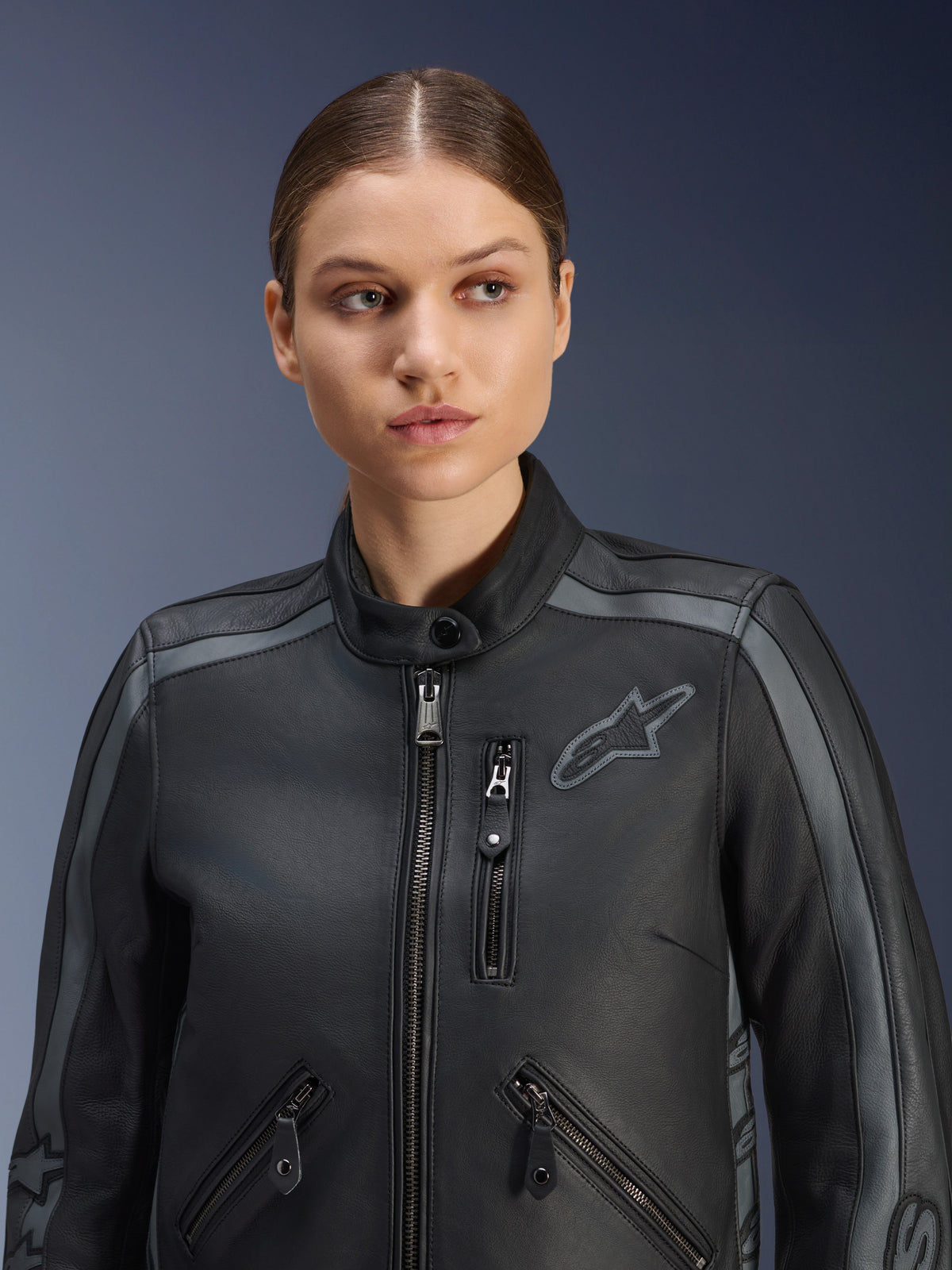 Kvinnlig modell iförd Alpinestars Stella Dyno Leather Jacket i Black Tar Gray Dark Gray, premium motorcykeljacka i nötlärder med heritage-design, grå axeldetaljer och flera fickor med dragkedja, skräddarsydd för en kvinnospecifik passform.