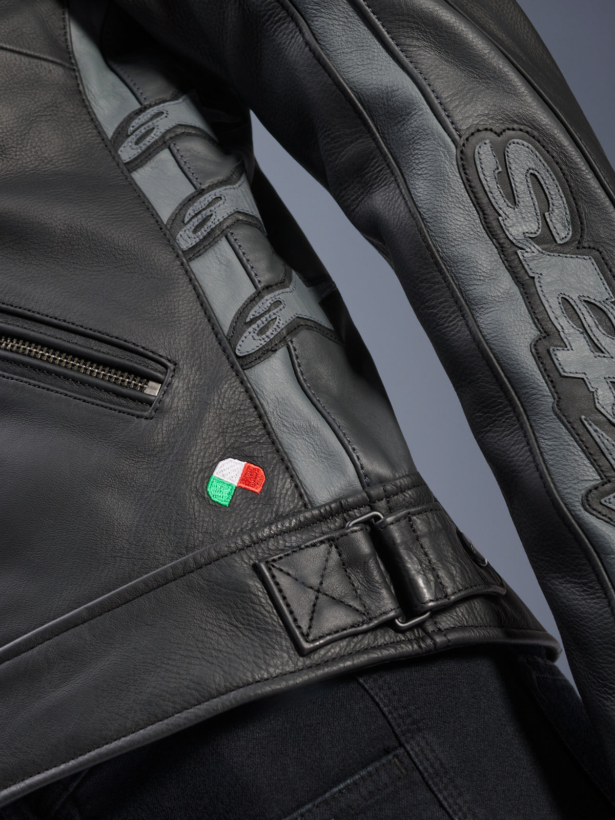 Alpinestars Stella Dyno skinnjacka, detalj, svart och Tar Gray nötläder, närbild med liten broderad italiensk flagga, justerbart midjespänne och grå läderpaneler med präglade Alpinestars-logotyper och Stella-branding på ärmen.