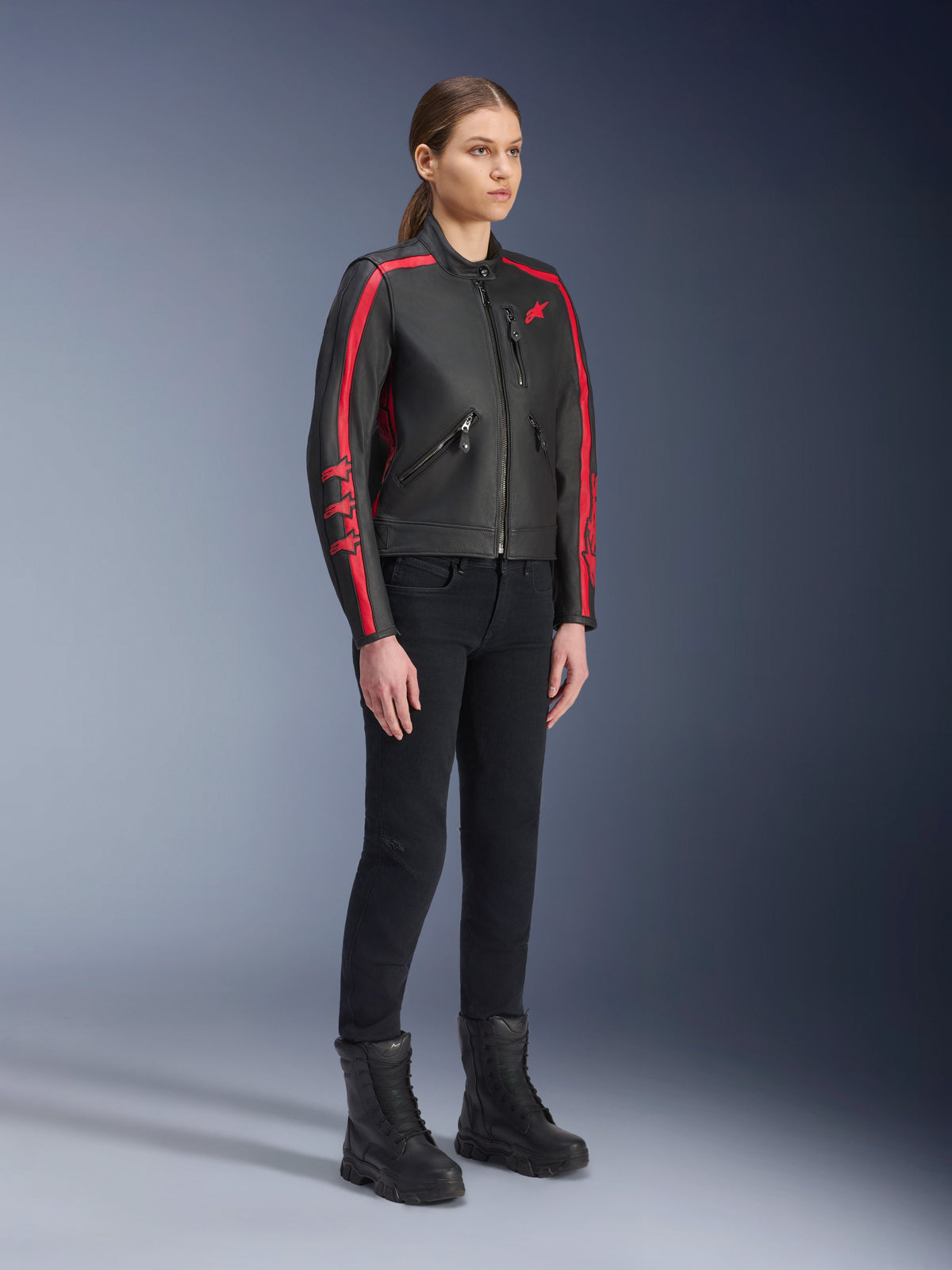 Kvinnlig modell som bär Alpinestars Stella Dyno läderjacka i Black Haute Red, matchad med svarta byxor och kängor, läderjacka för motorcykel i heritage-stil med röda ränder på ärmarna och röd logotyp, urban mc-utrustning för dam